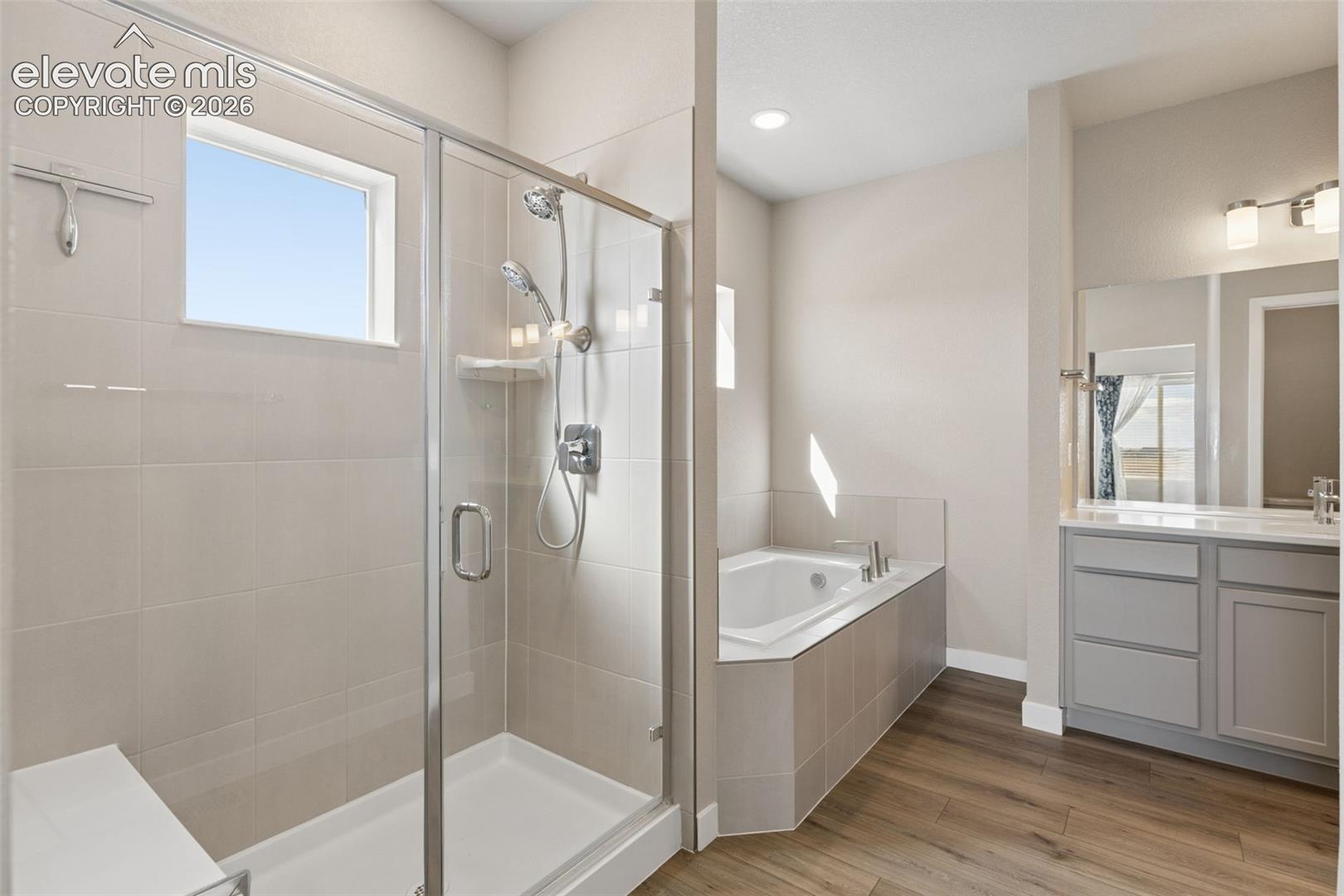 Bathroom featuring a glass-enclosed shower with a built-in bench, a separate soaking tub with tile surround, a vanity with neutral-toned cabinetry, and wood-finish flooring