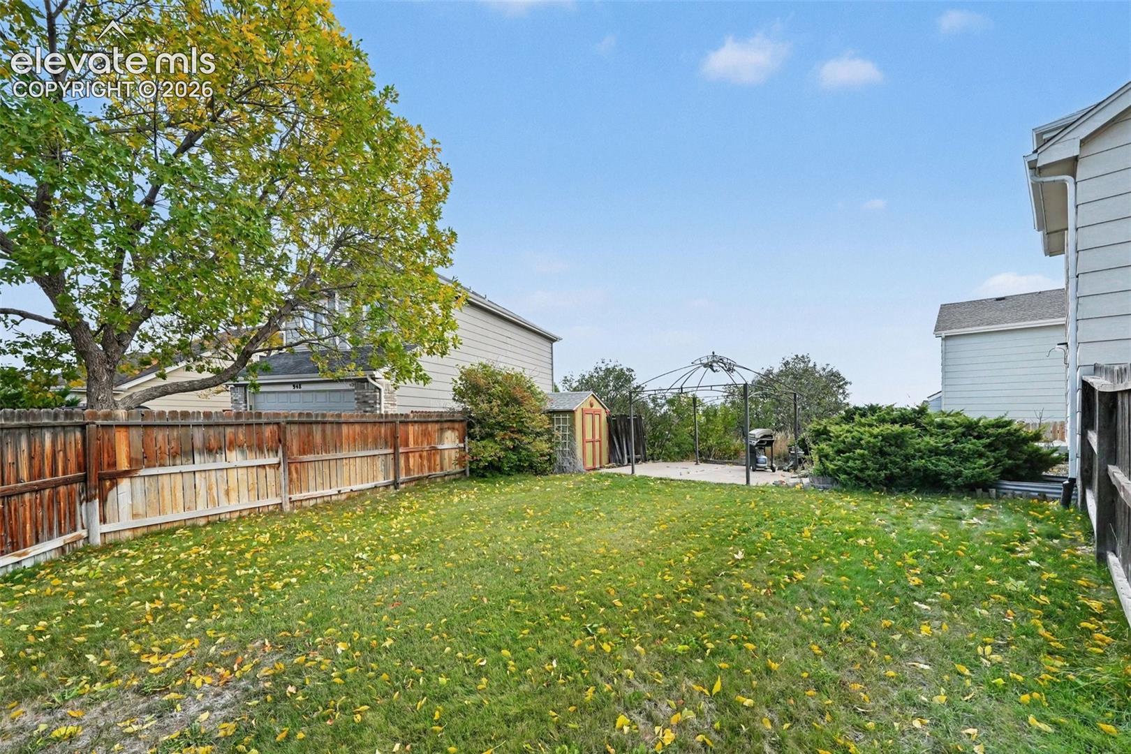 Fenced backyard featuring a grass lawn, mature trees, a storage shed, and a metal-framed gazebo