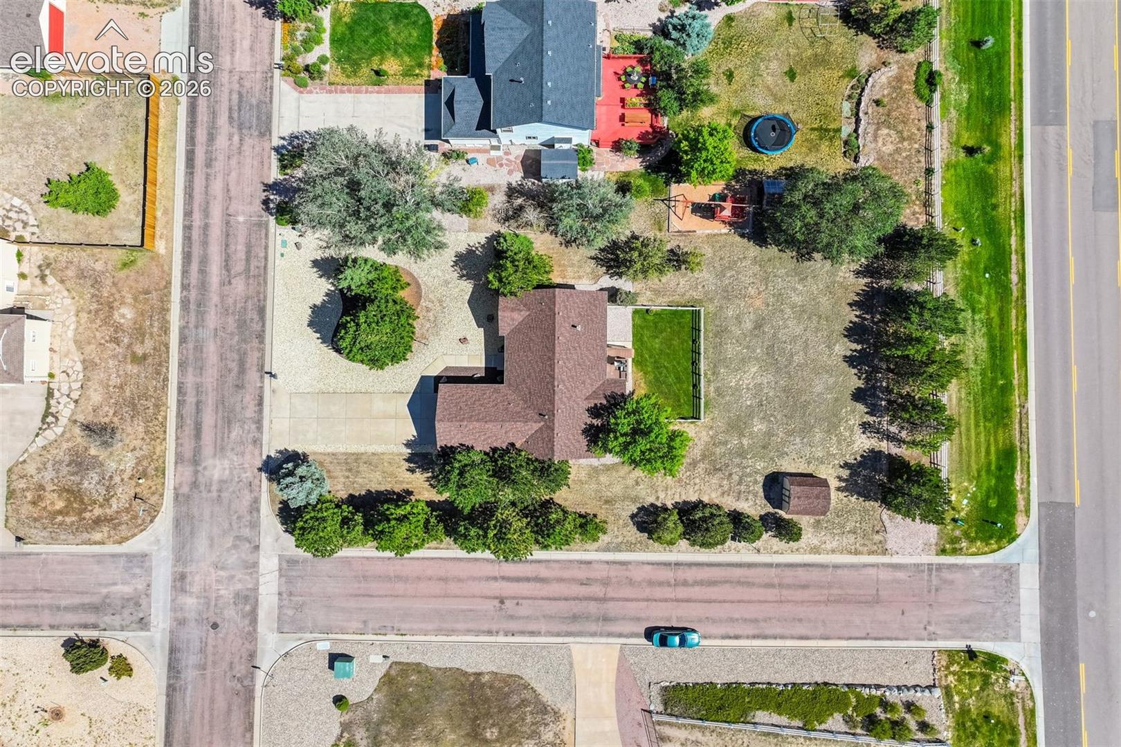 Aerial view of a corner lot property featuring mature landscaping, an expansive grass area, a concrete driveway, and an exterior storage structure