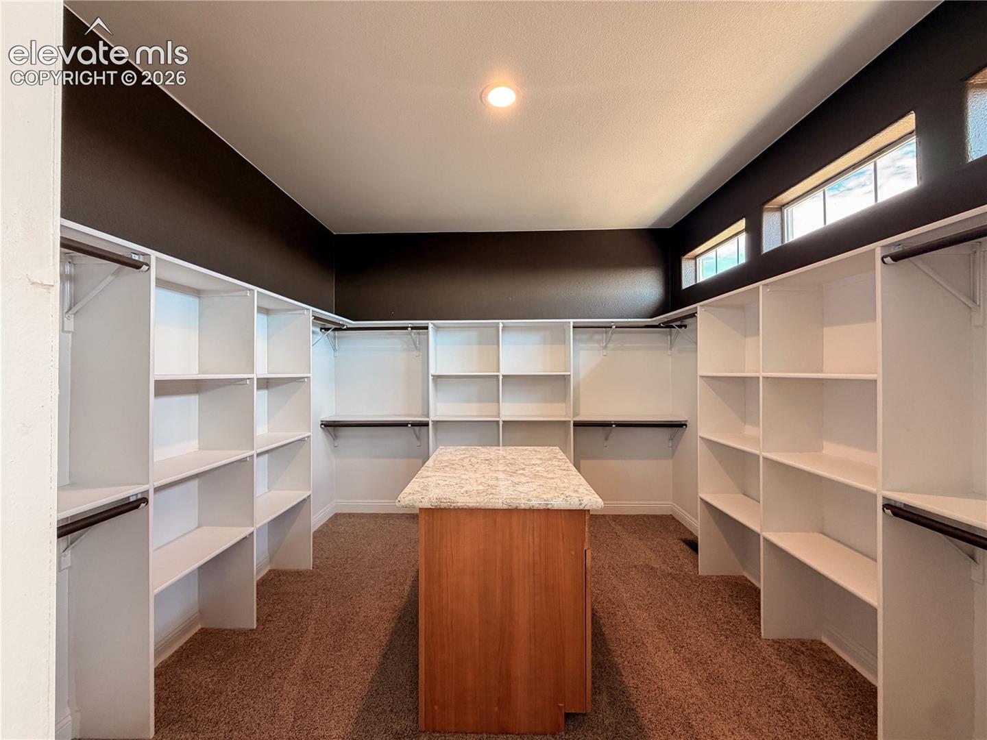 Walk-in closet featuring extensive built-in white shelving, a center island with a stone-finish countertop, high-reach clerestory windows, and recessed lighting
