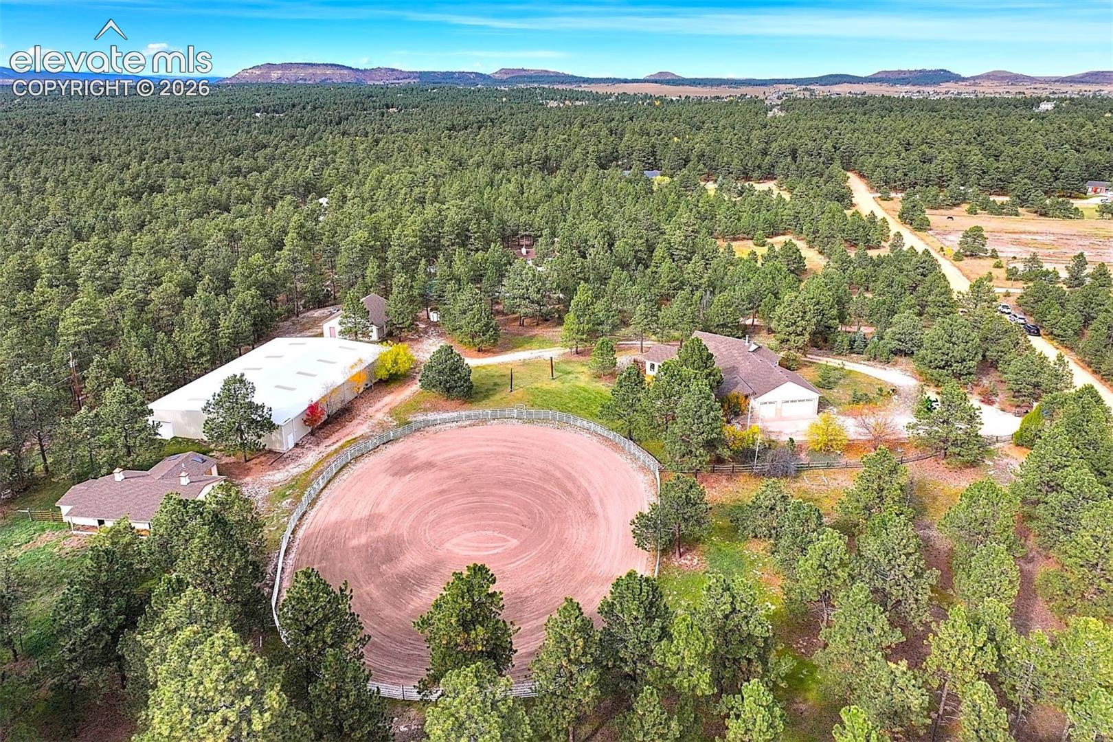 Aerial view of a sprawling property featuring multiple structures, a circular fenced riding arena, and extensive surrounding forest landscape