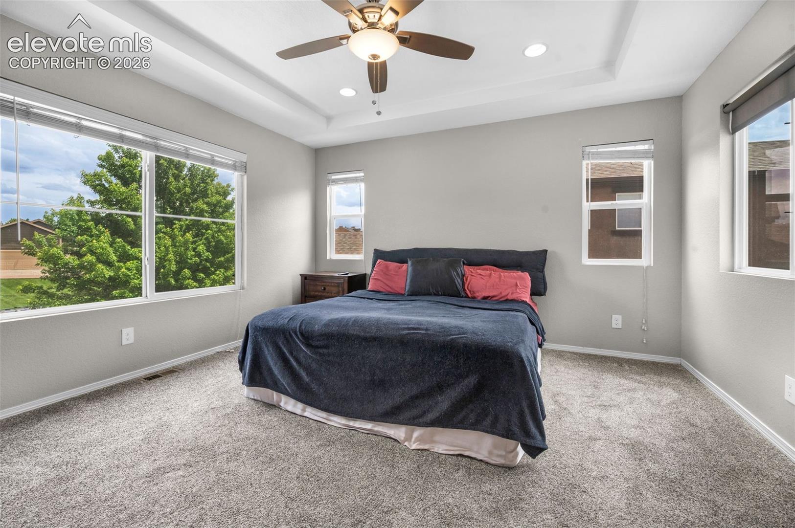 Primary bedroom featuring a tray ceiling with a ceiling fan and recessed lighting, multiple windows, and light gray wall paint