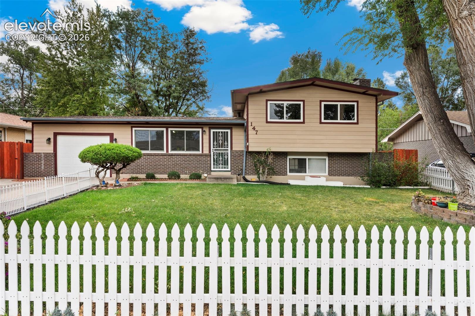 Split-level residence featuring a white picket fence, a manicured lawn, and mature trees