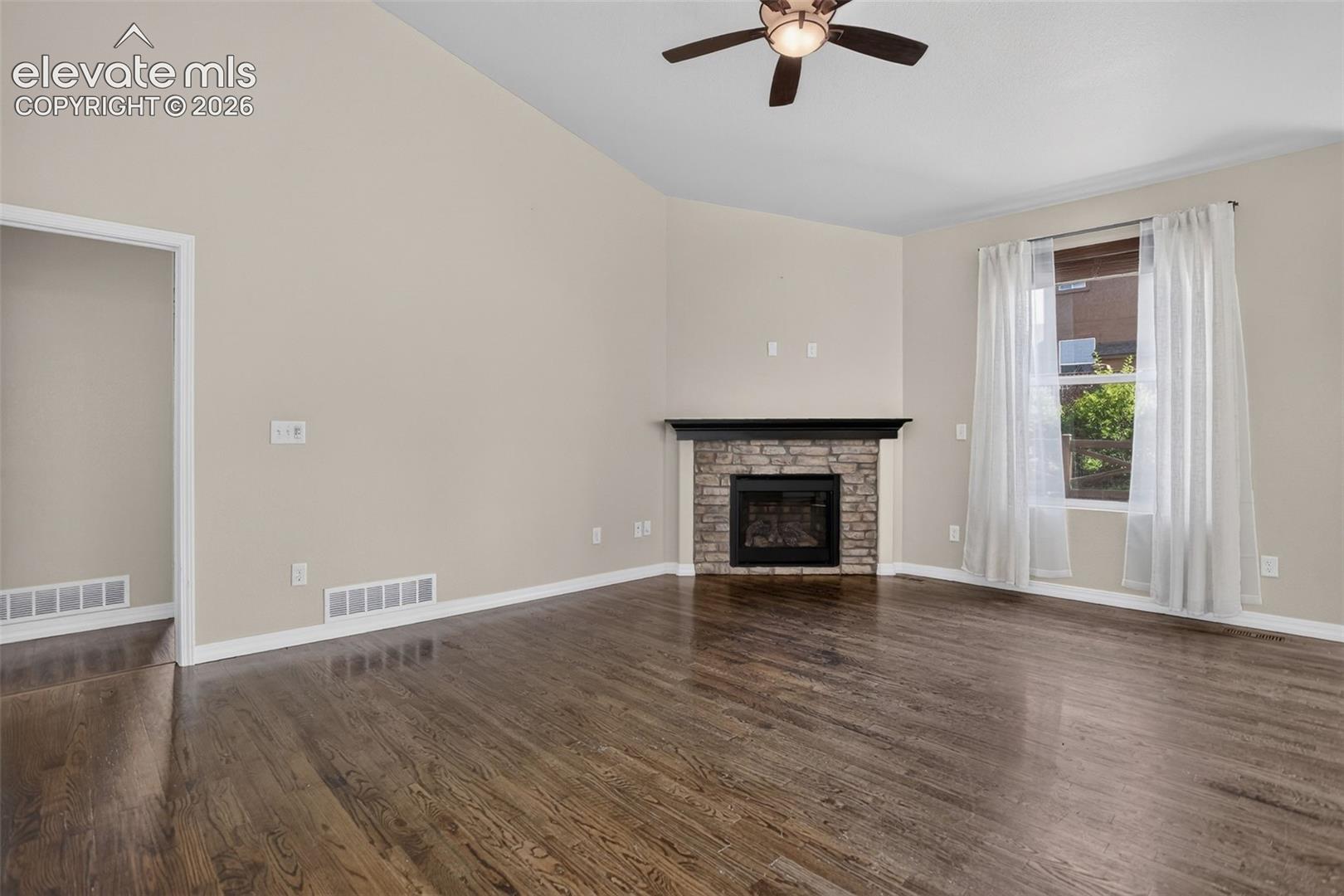 Vaulted ceiling, wood-finish flooring, corner fireplace with stone surround, window with white trim, and ceiling fan