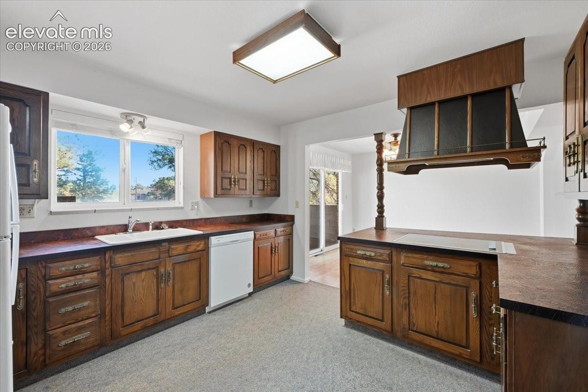 Kitchen featuring wood cabinetry, a custom wooden range hood, dark countertops, a double-basin sink, and an integrated cooktop