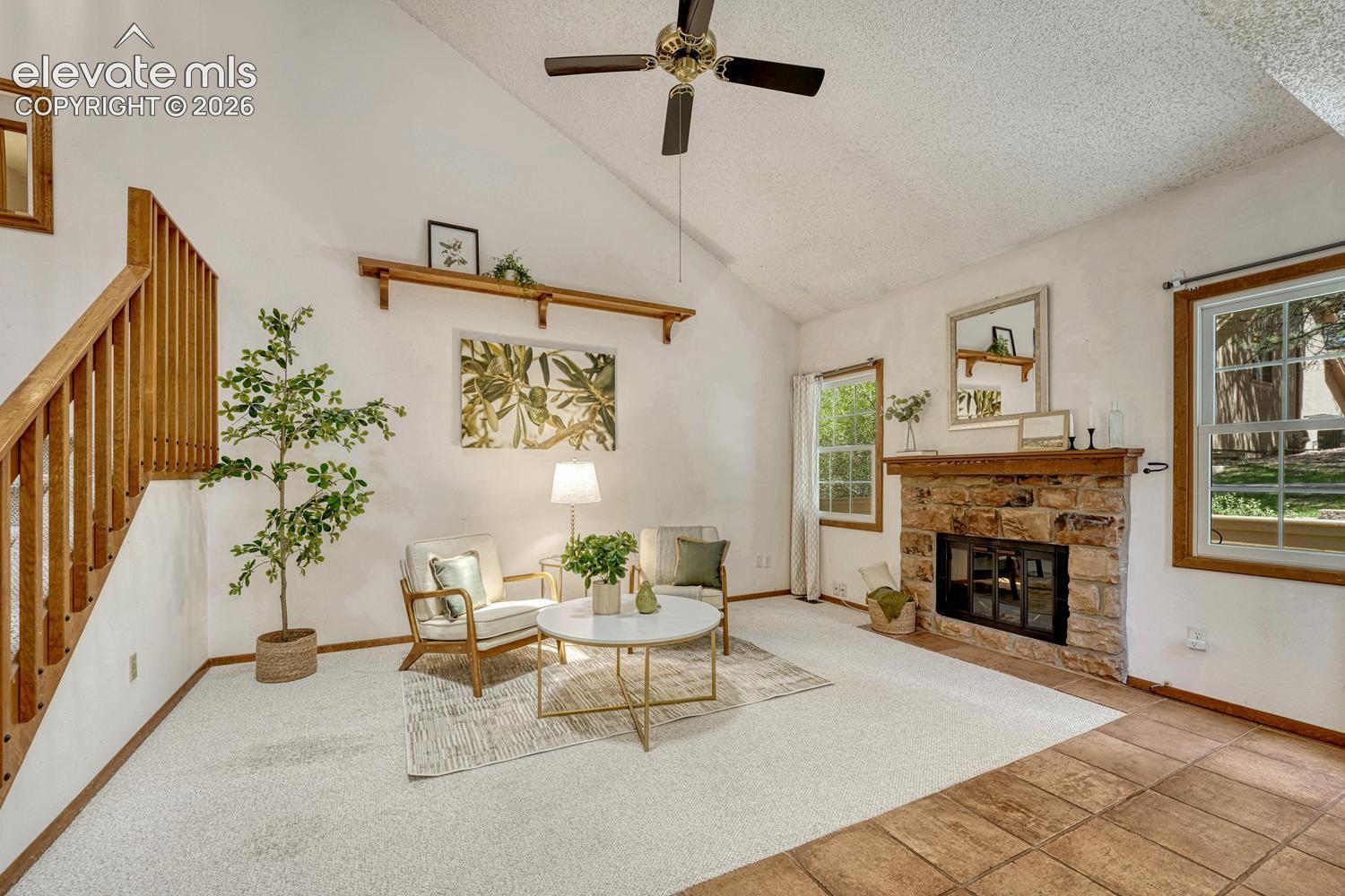 Bright, inviting vaulted living room featuring neutral carpeting, a ceiling fan, wall shelf, & cozy wood-burning fireplace with a classic brick surround to cozy up on chilly evenings. 