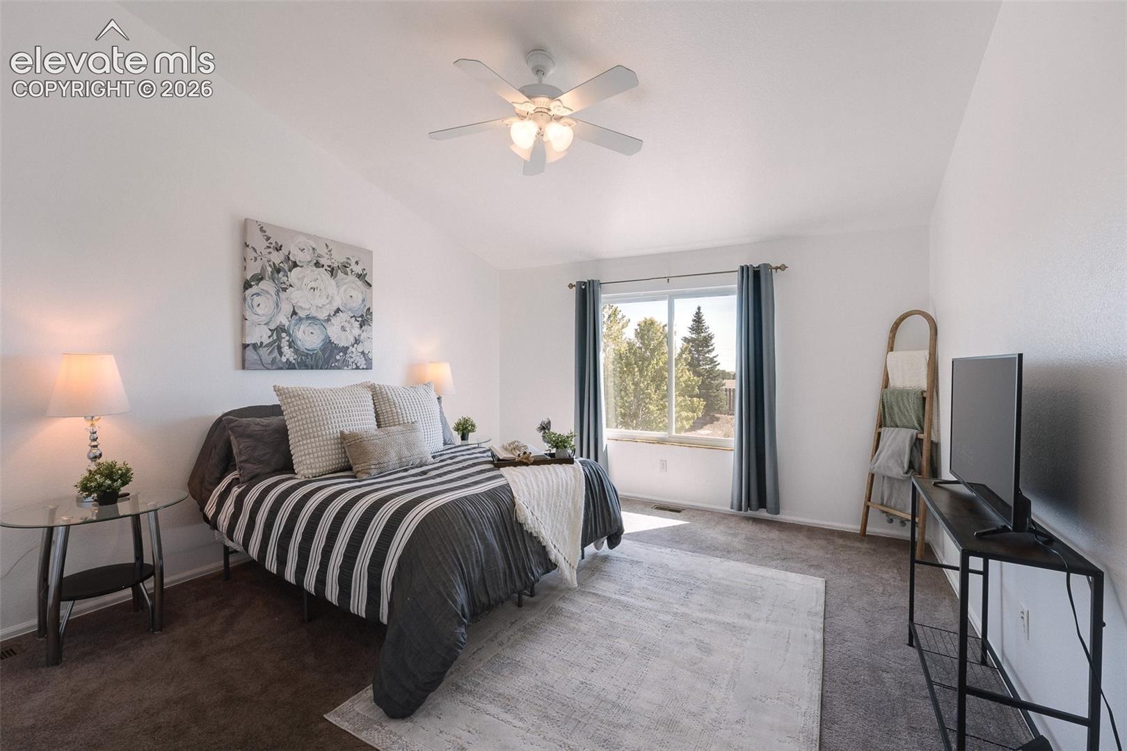The vaulted Primary Bedroom is a serene retreat with a lighted ceiling fan, neutral carpet, backyard views, a walk in closet, & adjoining 5-piece bathroom.