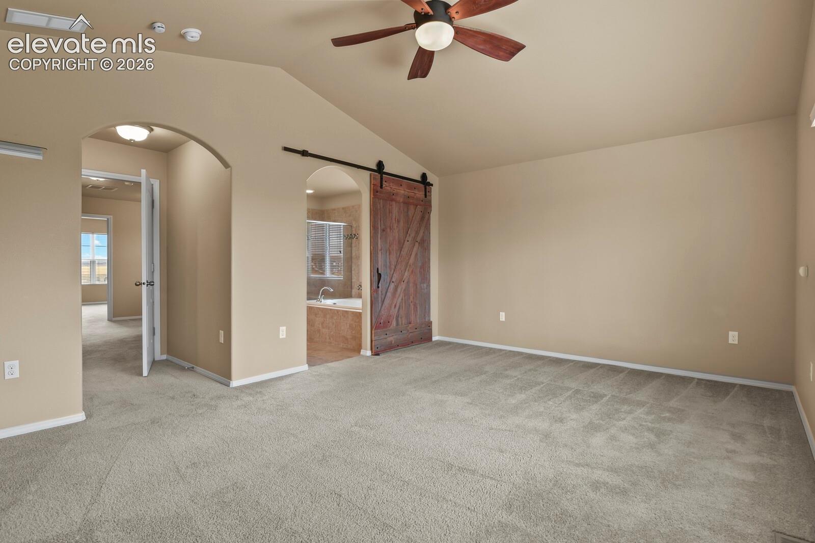 Vaulted ceiling, ceiling fan, plush carpeting, arched doorways, and a sliding barn door