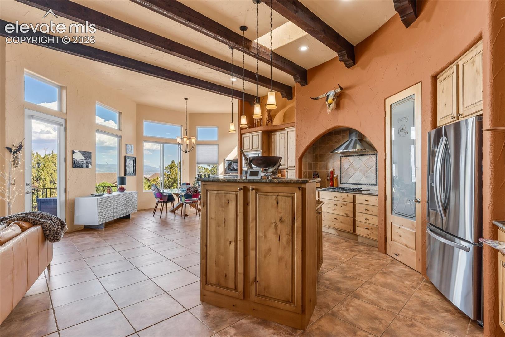 Open-concept kitchen featuring stained wood cabinetry, stone-tiled flooring, an arched cooking alcove, and exposed timber ceiling beams