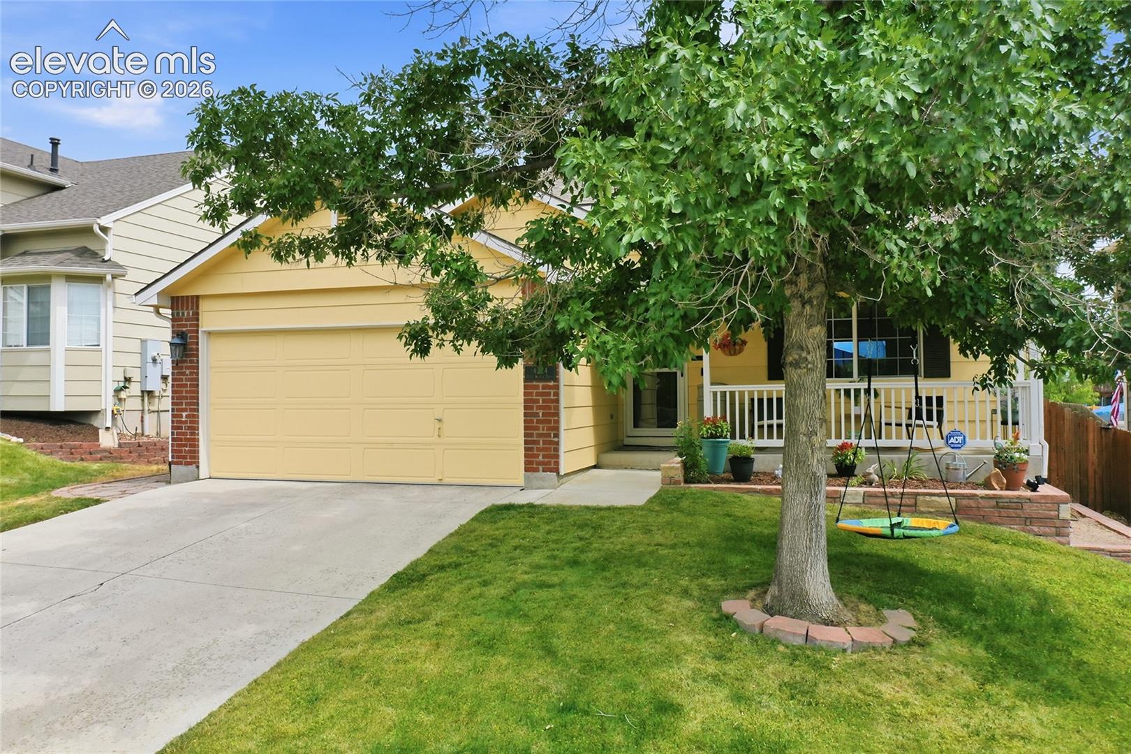 Single-story residence featuring light yellow siding, brick exterior accents, an attached two-car garage, a covered front porch with white railing, and a concrete driveway