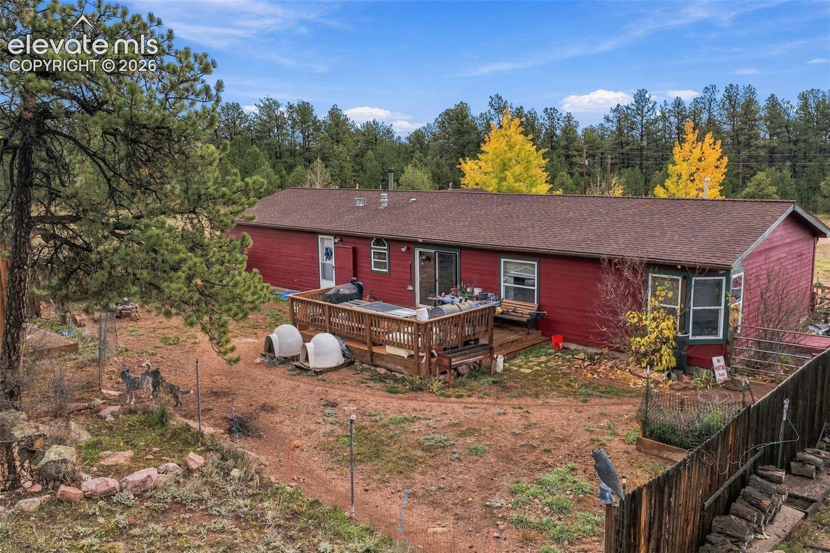 Single-story residence featuring red wood siding, a composition shingle roof, a wood deck, and a multi-pane bay window, situated on a lot with mature pine trees