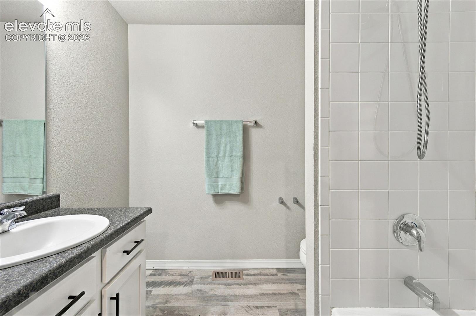 Bathroom featuring white cabinetry, dark stone-finish countertop with an integrated basin, wood-finish flooring, and white square-tile shower surround with a wall-mounted shower head