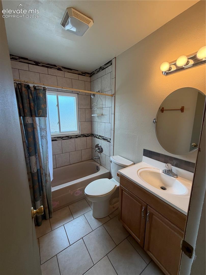 Full bathroom featuring a wood-finish vanity with a drop-in sink, circular wall-mounted mirror, tiled shower enclosure with decorative border inlay, and ceramic tile flooring