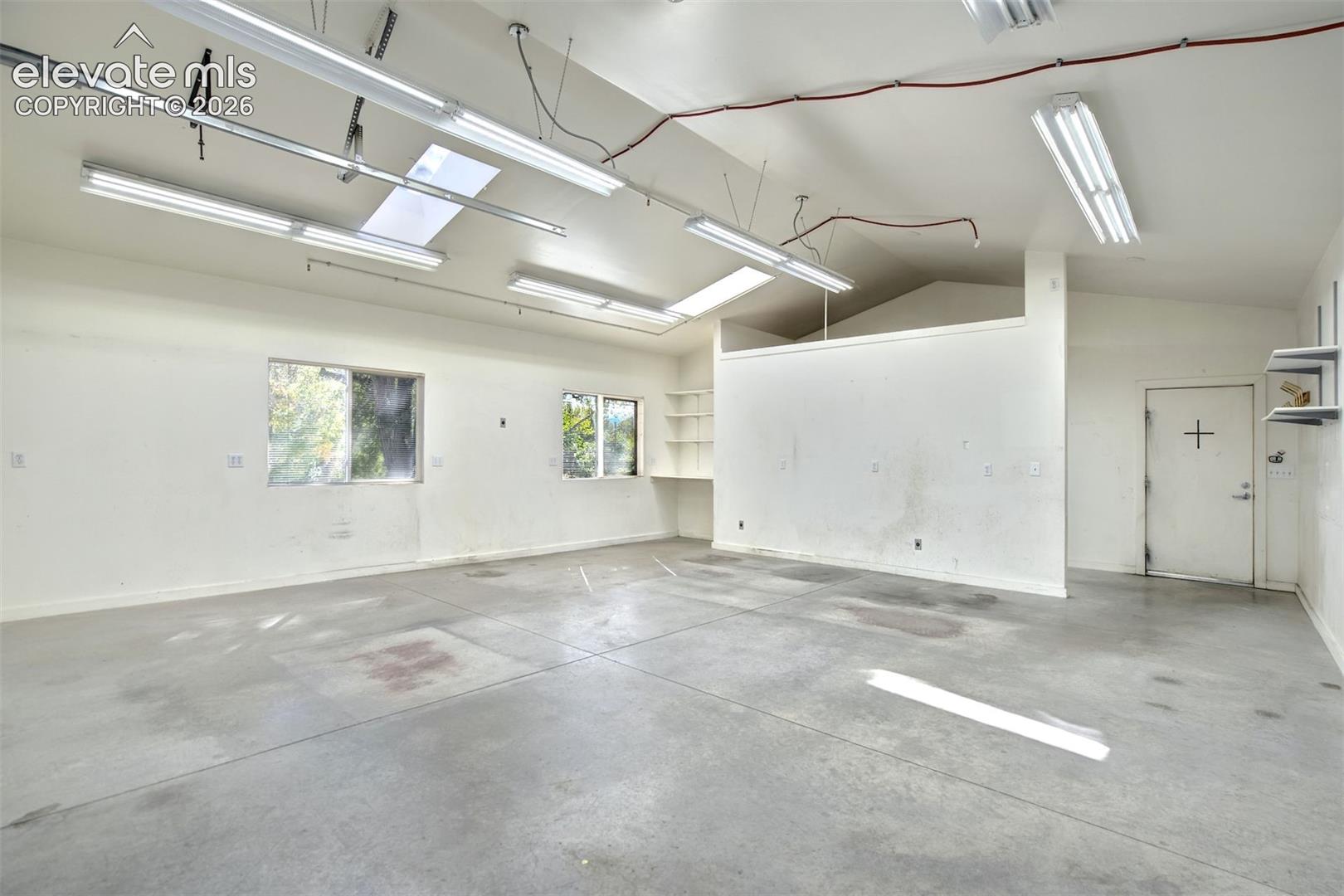 Spacious workshop area featuring high vaulted ceilings, multiple linear fluorescent light fixtures, polished concrete flooring, built-in wall shelving, and dual windows