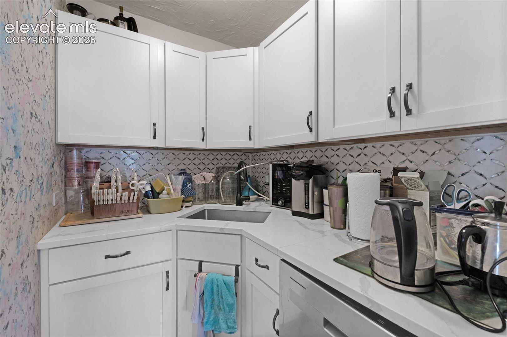 White shaker-style cabinetry with dark metal pulls, stone-finish countertops, patterned tile backsplash, and stainless steel under-mount sink with matte black faucet