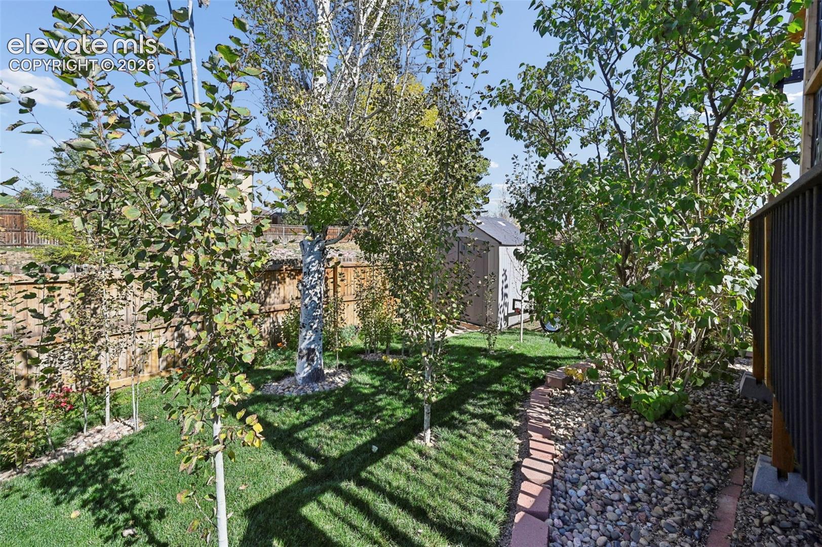 Rear yard featuring a manicured lawn, established trees, a fenced perimeter, stone-bordered landscaping beds, and a detached storage structure