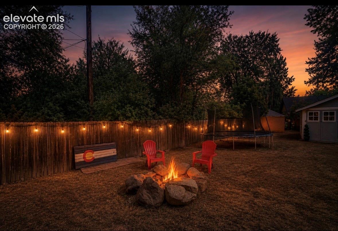 Private backyard featuring a rock fire pit, wood fence, string lighting, and mature perimeter trees