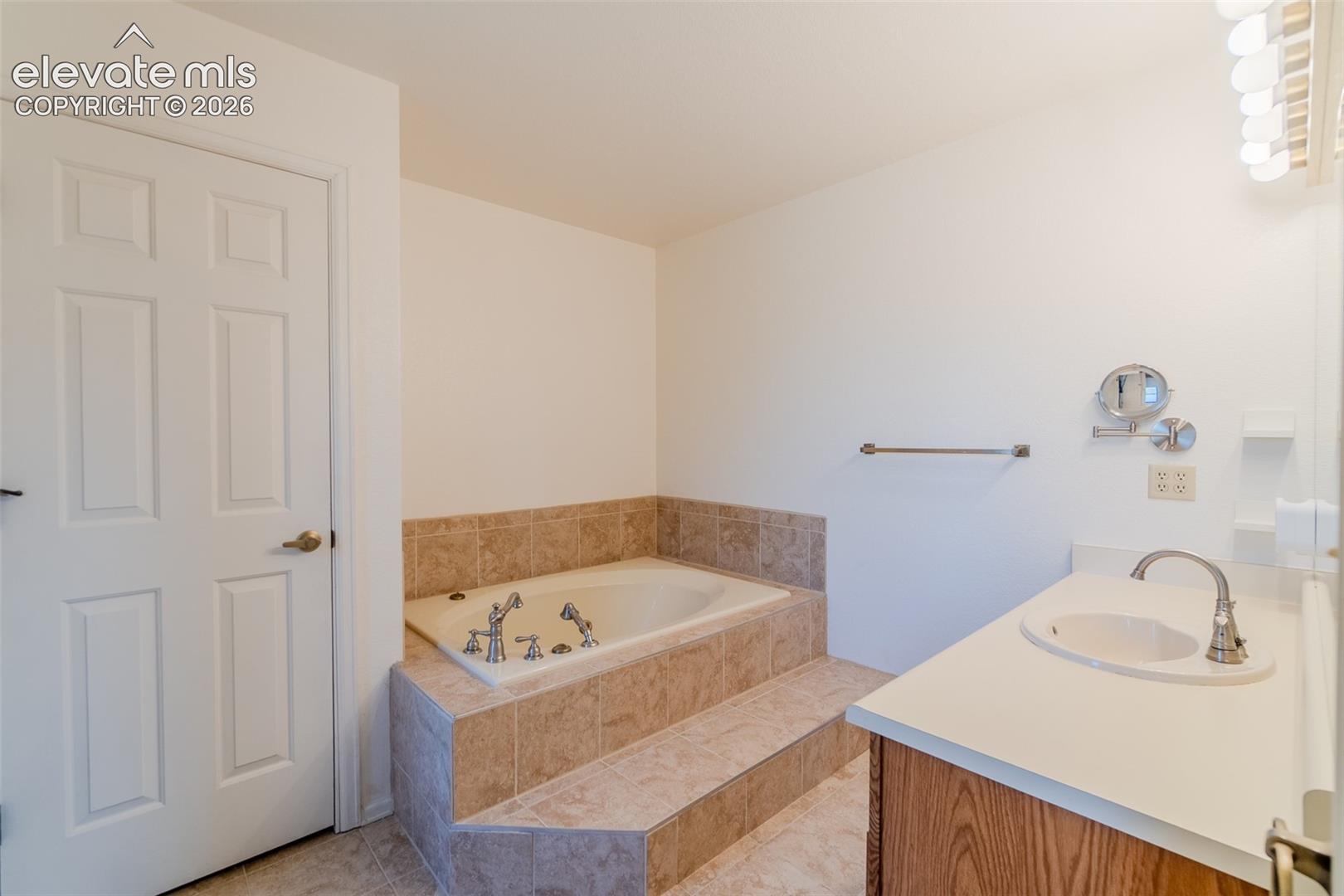 Built-in jetted tub with tile surround, wood-finish vanity with countertop sink, and wall-mounted vanity lighting