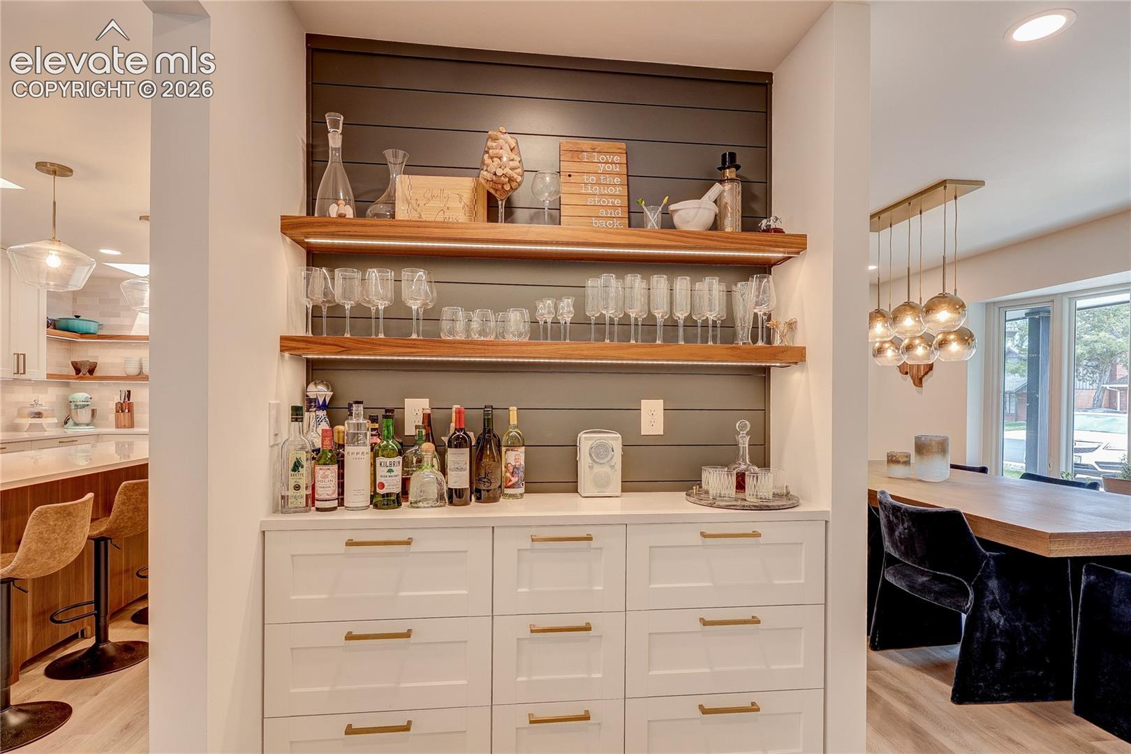 Built-in wet bar with white cabinetry, gold-finish hardware, open wood shelving, and a shiplap accent wall