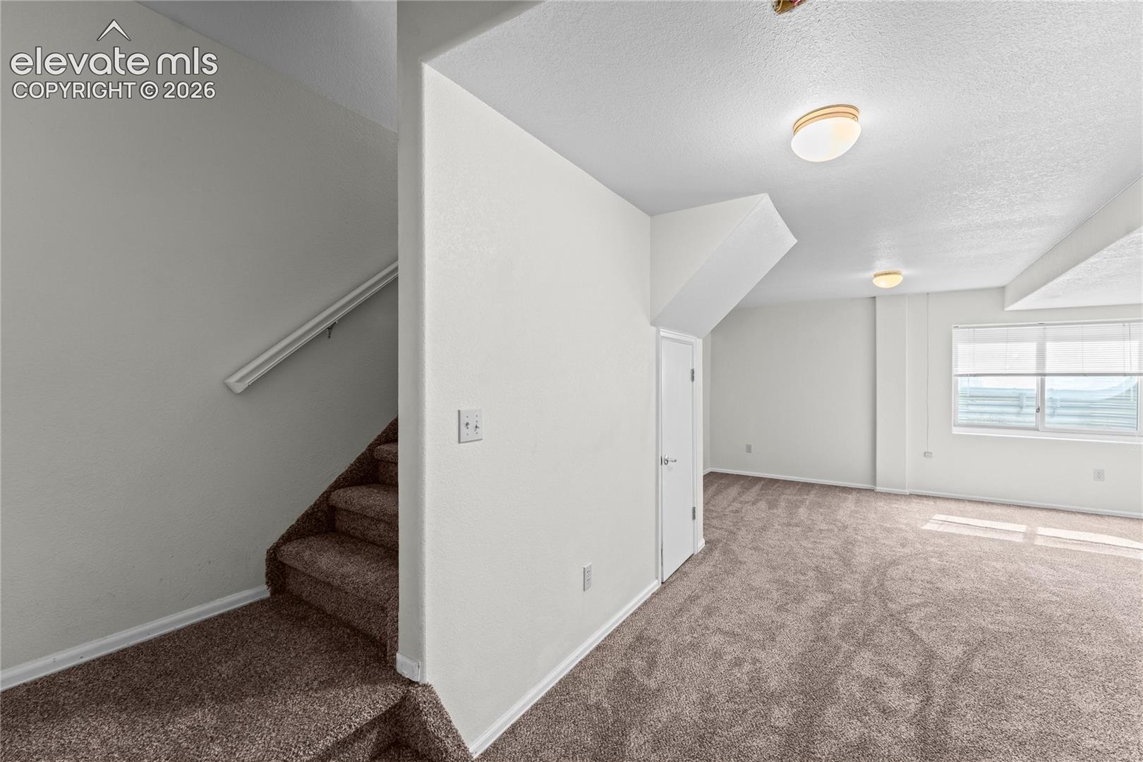 Carpeted stairs with handrail leading to an open room featuring carpeted flooring, white walls, and a window with blinds