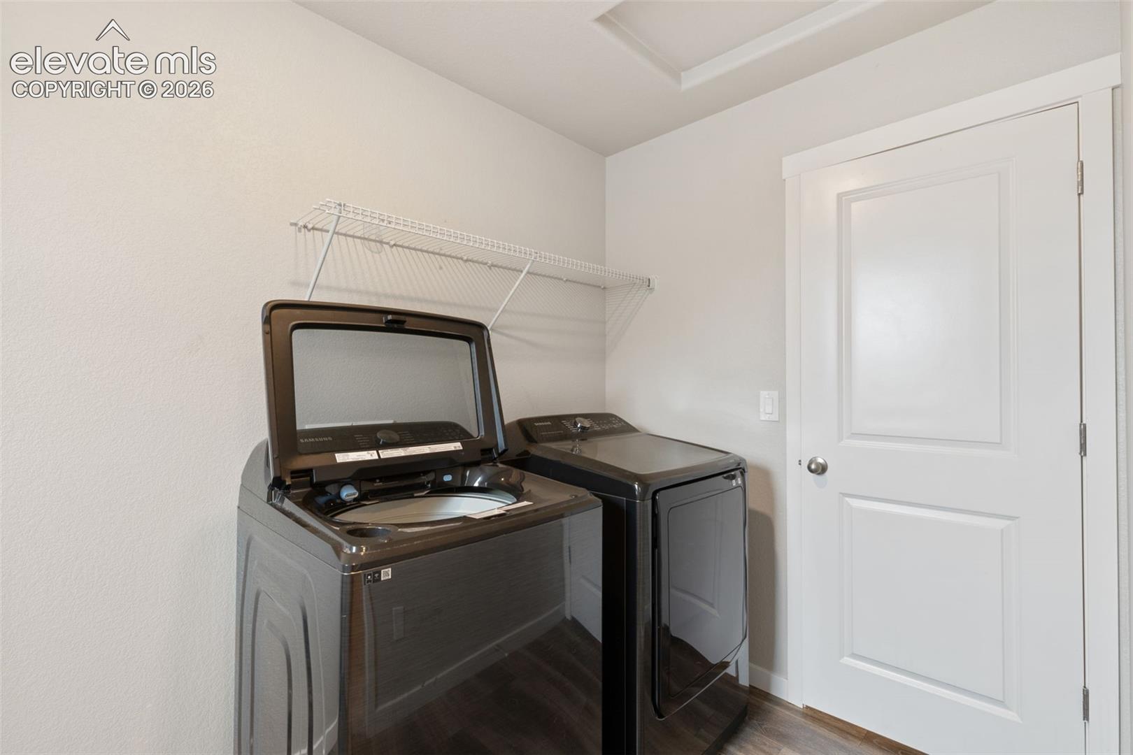 Dedicated laundry area featuring wood-finish flooring, a white interior door with panel detailing, and an overhead wire shelving unit