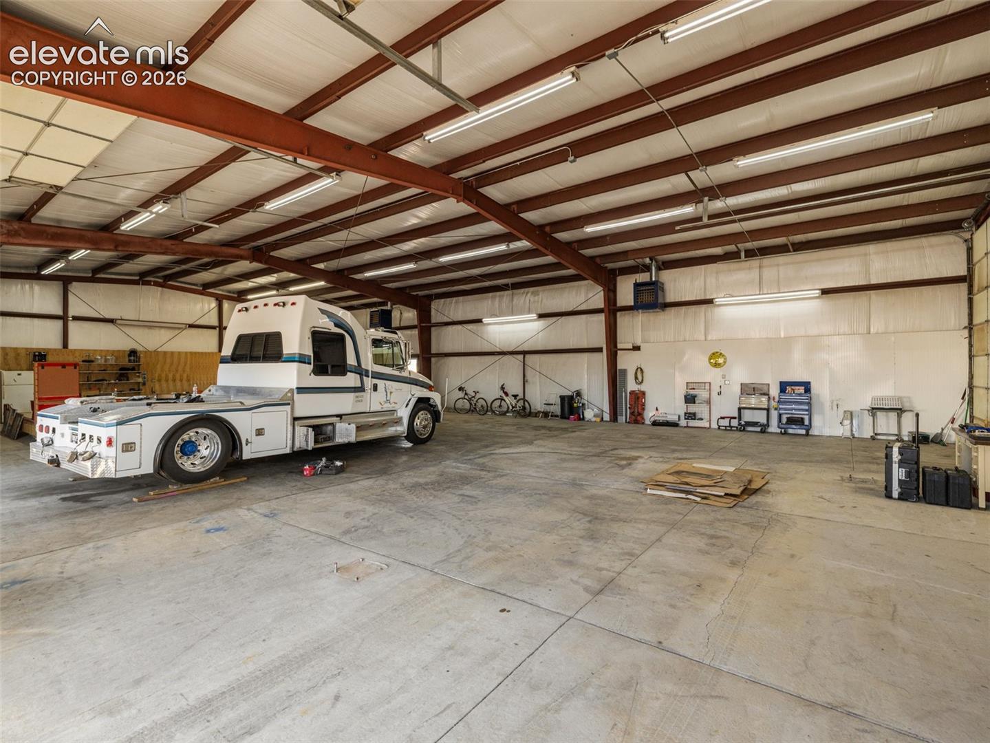 Expansive workshop featuring exposed steel beam framing, high-clearance ceilings, industrial-grade concrete flooring, and multiple linear fluorescent light fixtures