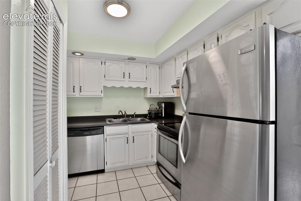 Kitchen featuring stainless steel appliances, white cabinetry, dark countertops, a double basin sink, and tile flooring