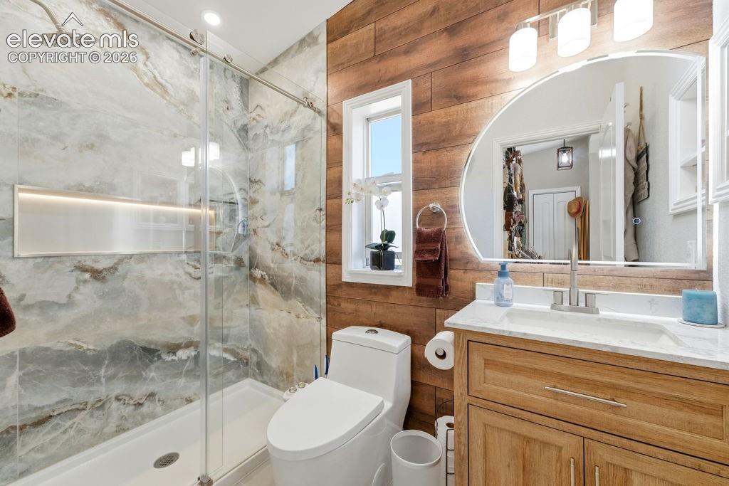 Walk-in shower with natural stone-inspired tile and integrated niche, wood-finish accent wall, single window, and a vanity with a white countertop and undermount sink