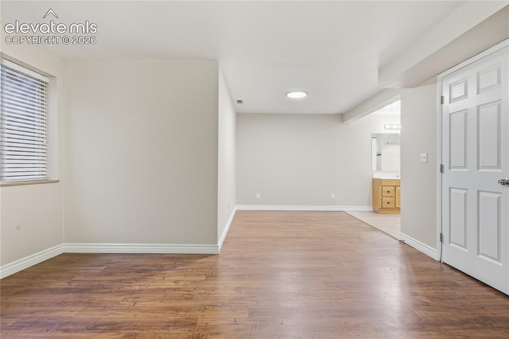 Spacious room featuring wood-finish flooring, a window with blinds, and white baseboards