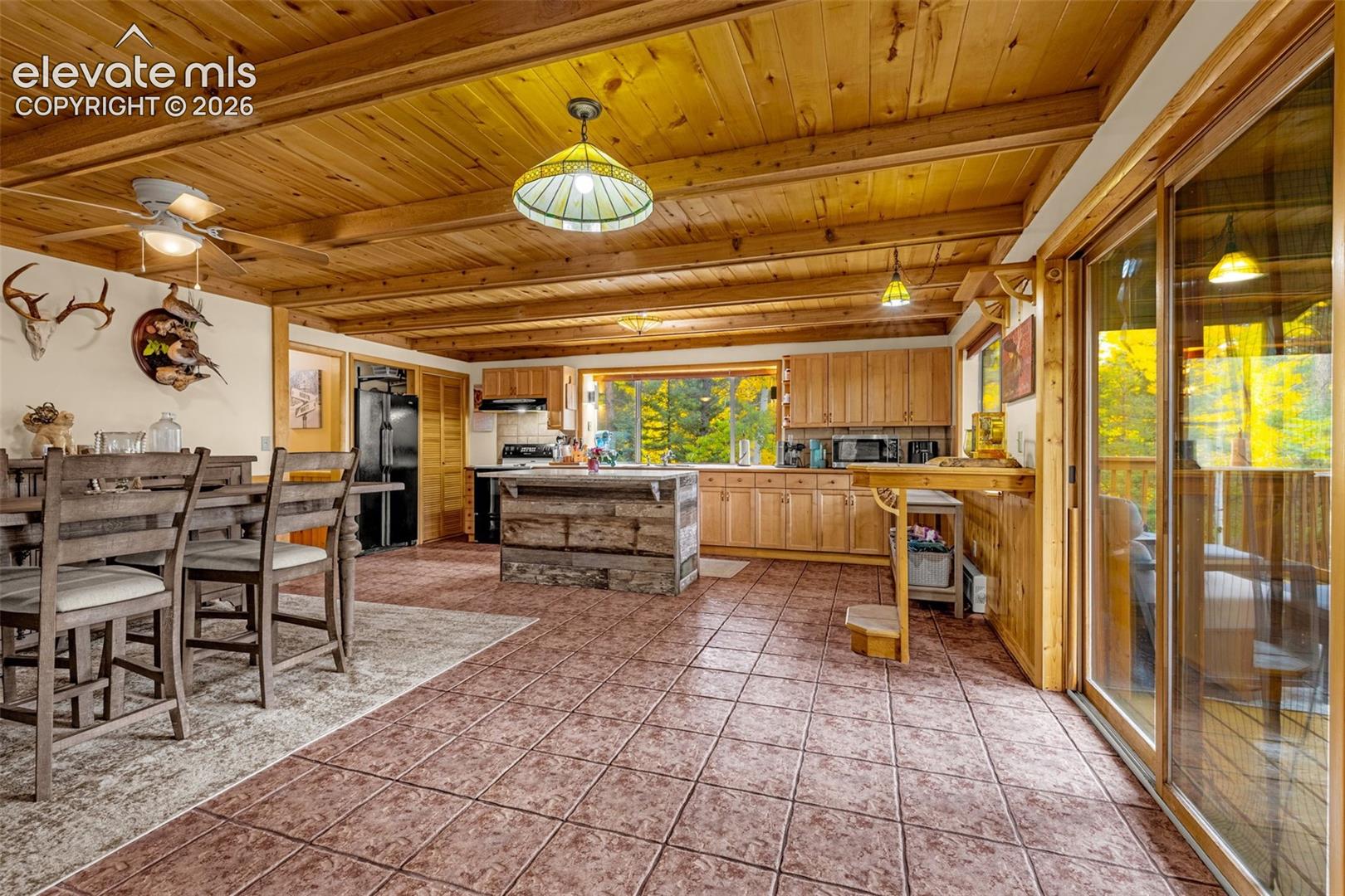 Exposed beam ceiling with wood-paneled finish, ceramic tile flooring, custom kitchen cabinetry, kitchen island with rustic wood detailing, and floor-to-ceiling glass sliding doors