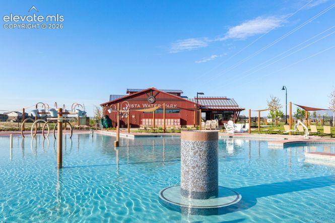 Recreational aquatic facility featuring a large swimming pool, integrated water play structures, a central tile-finished pillar, and a multipurpose building with metal roofing