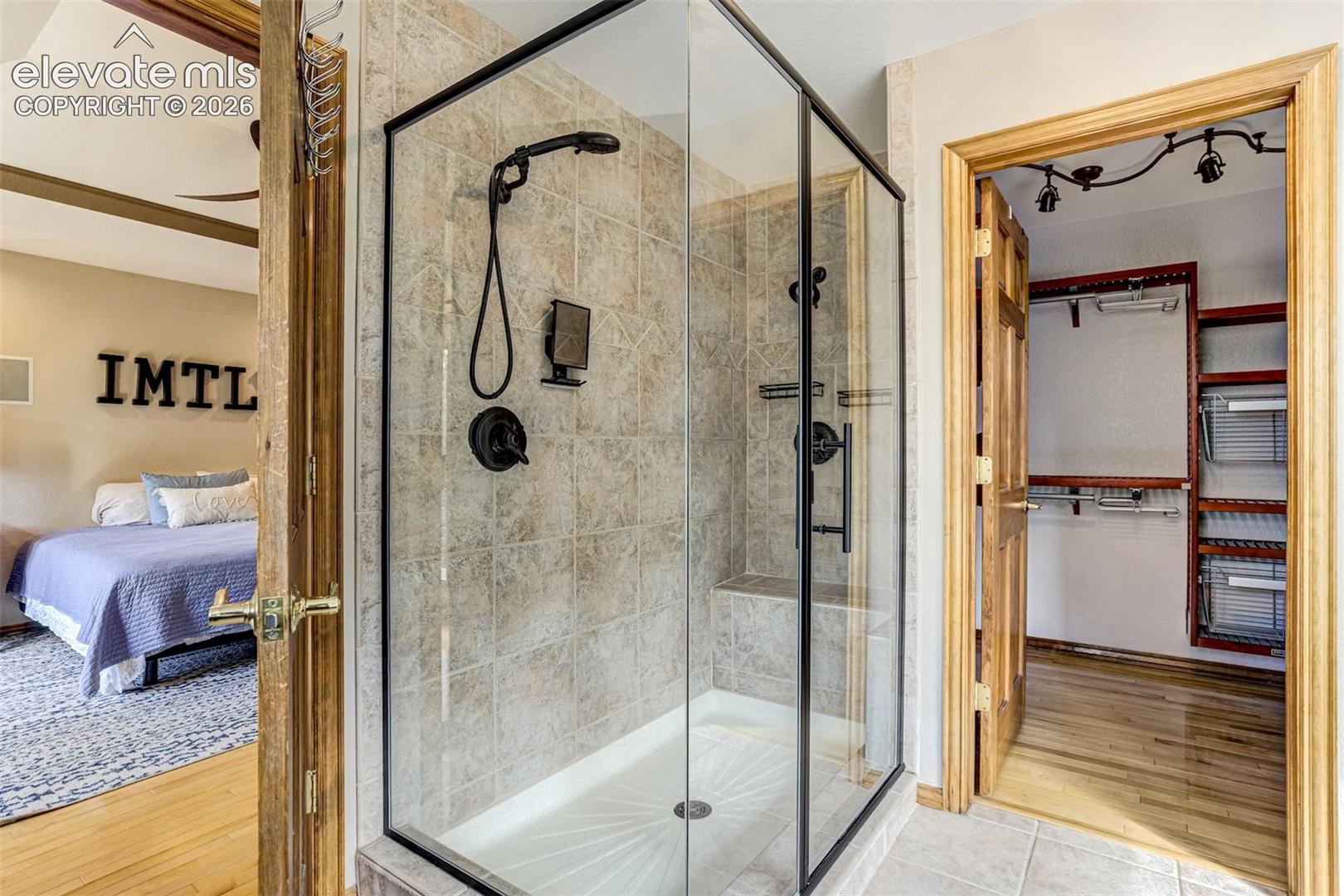 Primary Bathroom Suite Featuring a Frameless Glass Enclosure Shower with Dual Showerheads, Stone Tile Surround, Matte Black Hardware Fixtures, and Direct Access to a Walk-In Closet with Custom Built-In Shelving and Organizational Systems