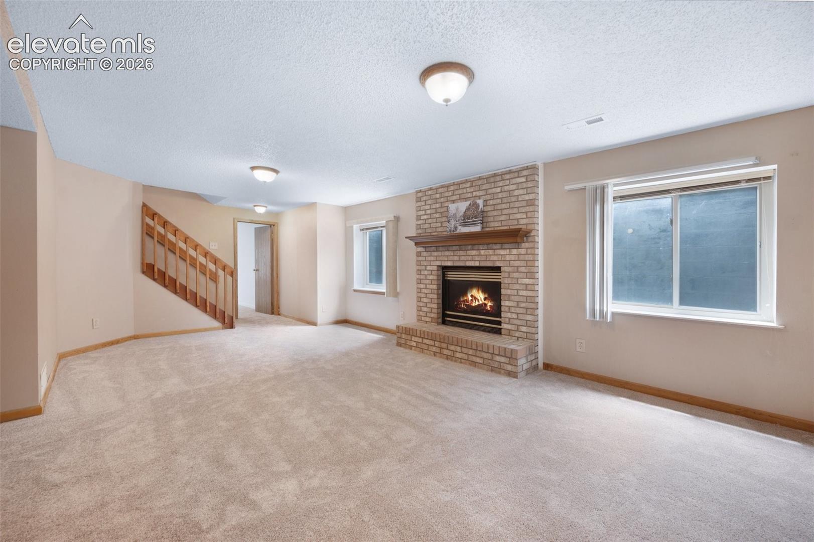 Unfurnished living room featuring a textured ceiling, a brick fireplace, healthy amount of natural light, stairs, and baseboards. fireplace staged and added.