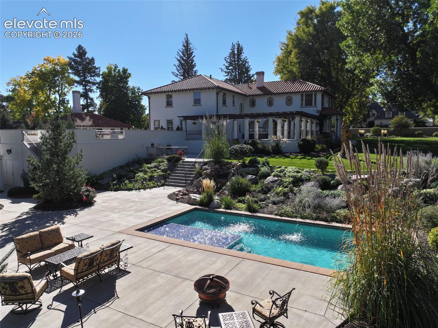 Stucco exterior featuring a clay tile roof, expansive concrete patio, built-in swimming pool with tiled steps, tiered rock garden, and a covered pergola