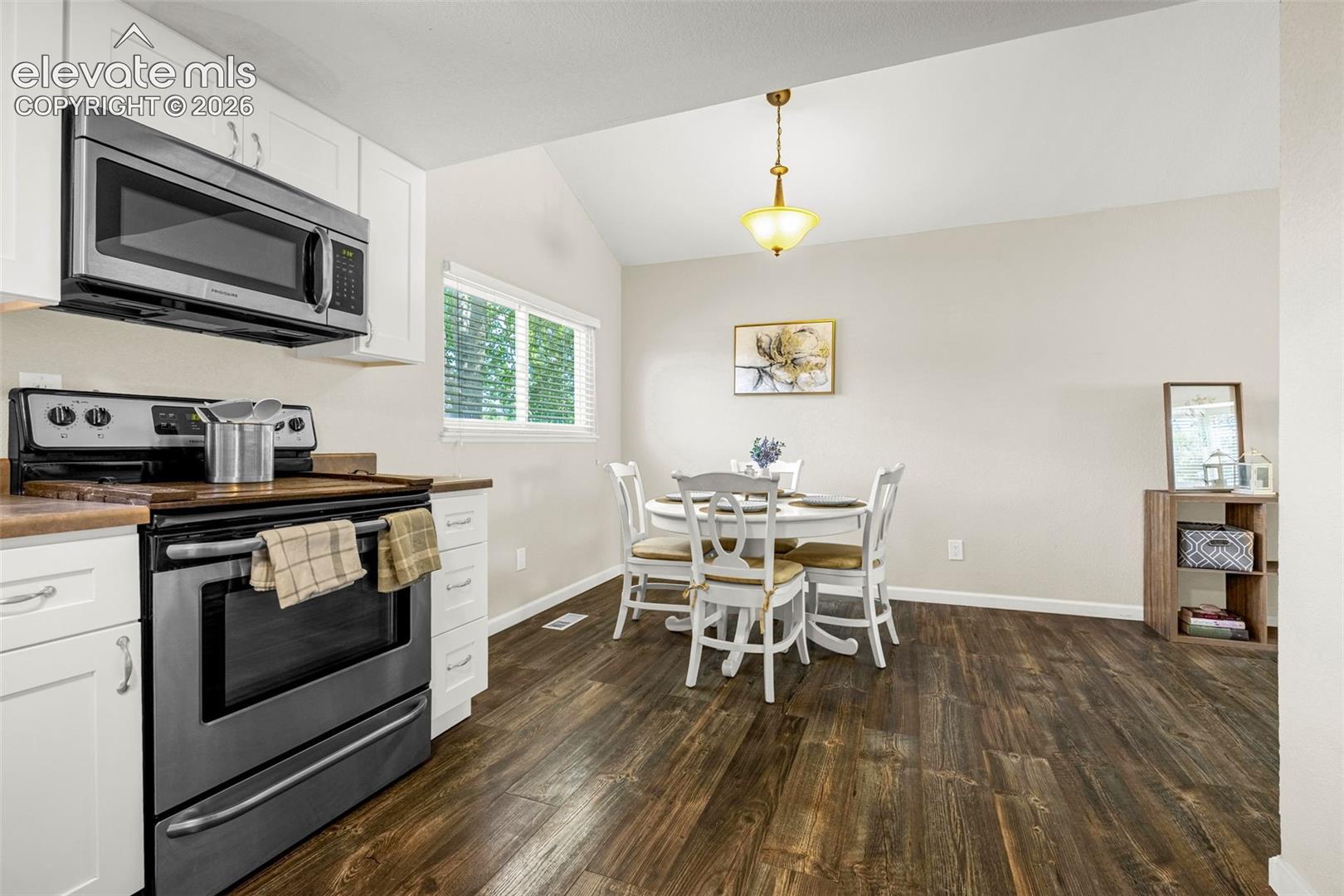 Kitchen and dining area featuring vaulted ceilings, wood-finish flooring, white cabinetry, stainless steel appliances, and a pendant light fixture