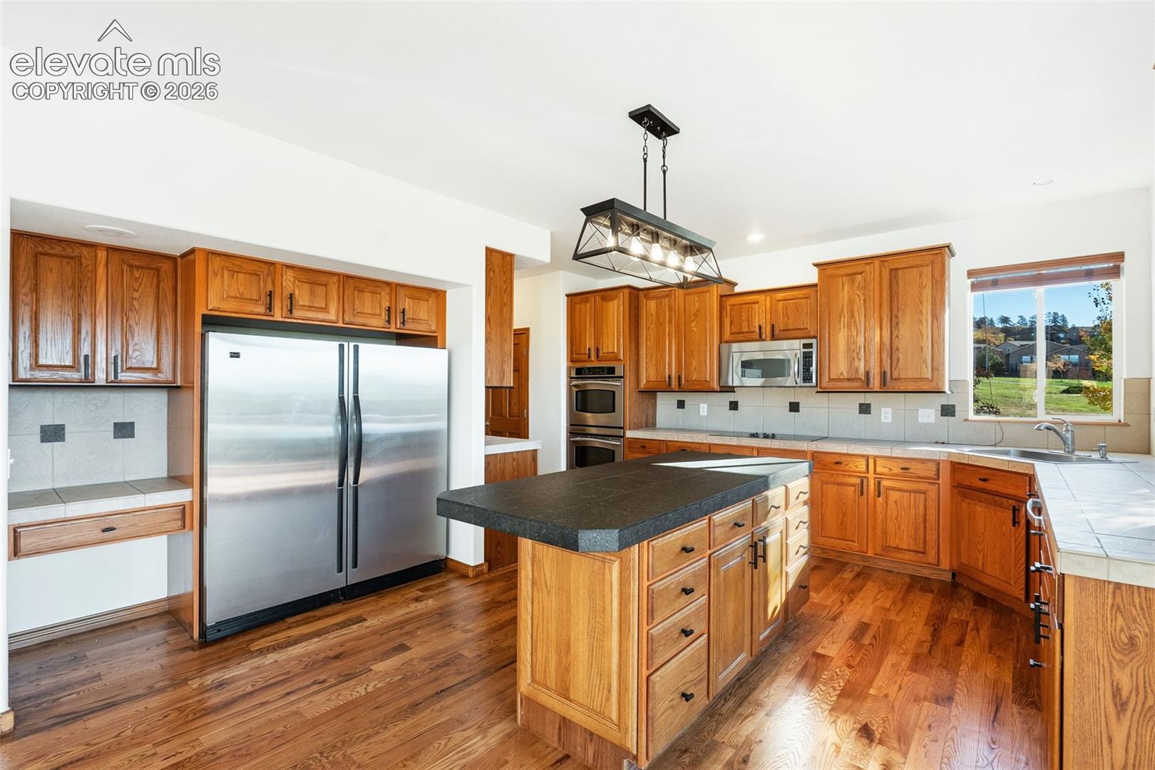 Kitchen featuring wood cabinetry, stainless steel appliances including a double wall oven and built-in microwave, a center island with a dark countertop, and wood-finish flooring