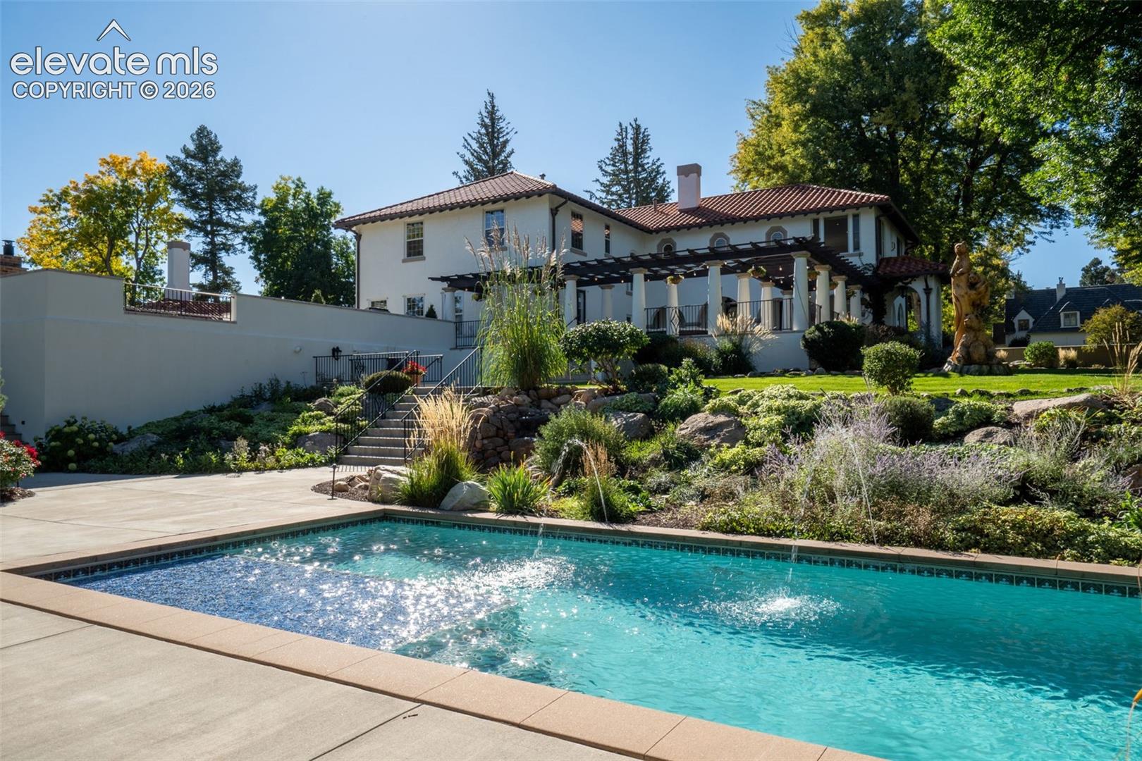 In-ground swimming pool featuring a tile border and water feature, concrete pool deck, covered pergola, stucco exterior, and multi-level stone landscaping