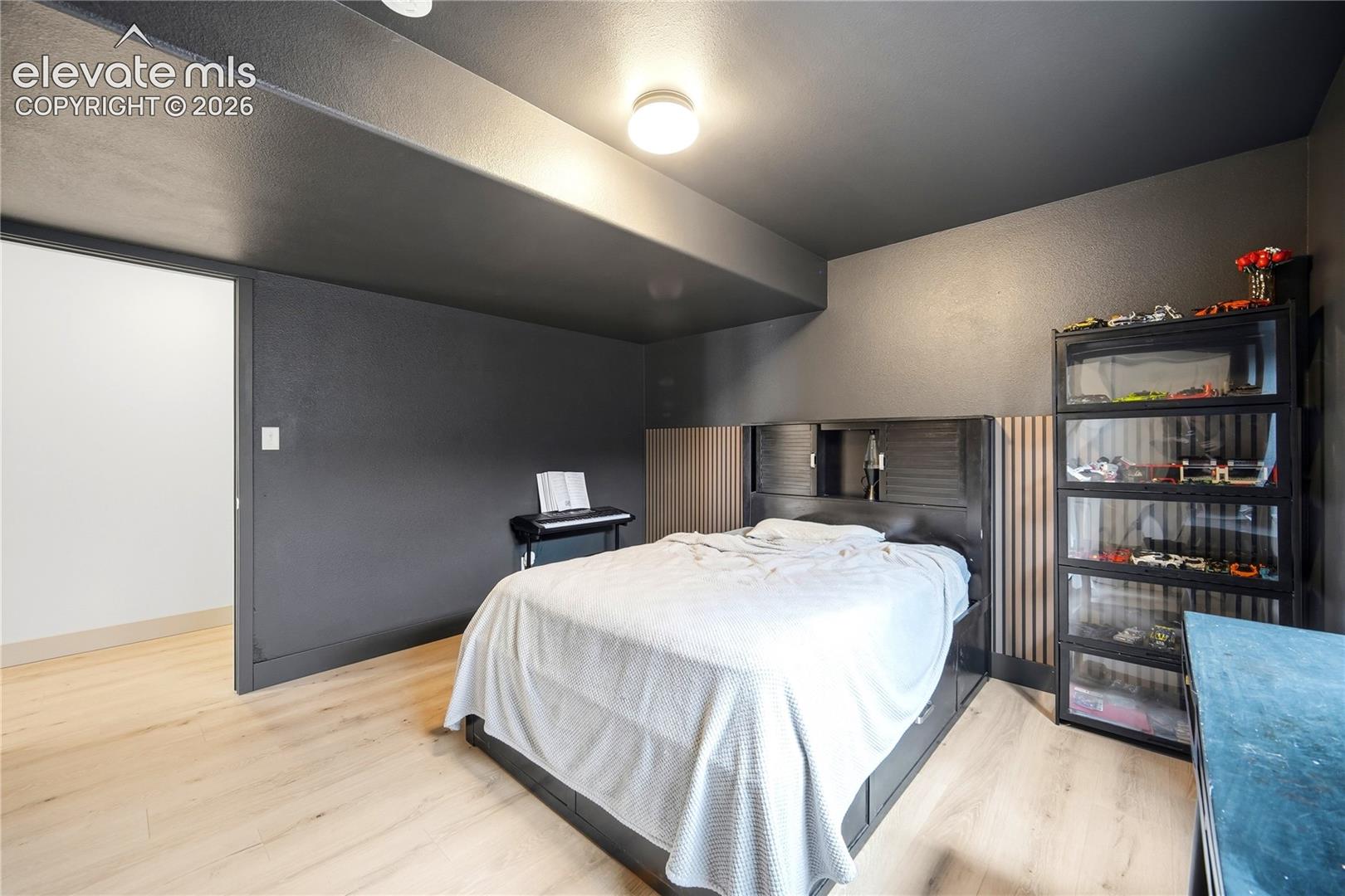 Room featuring light-toned wood-finish flooring, dark-toned walls, a built-in headboard with vertical slat accents, and a flush-mount ceiling light