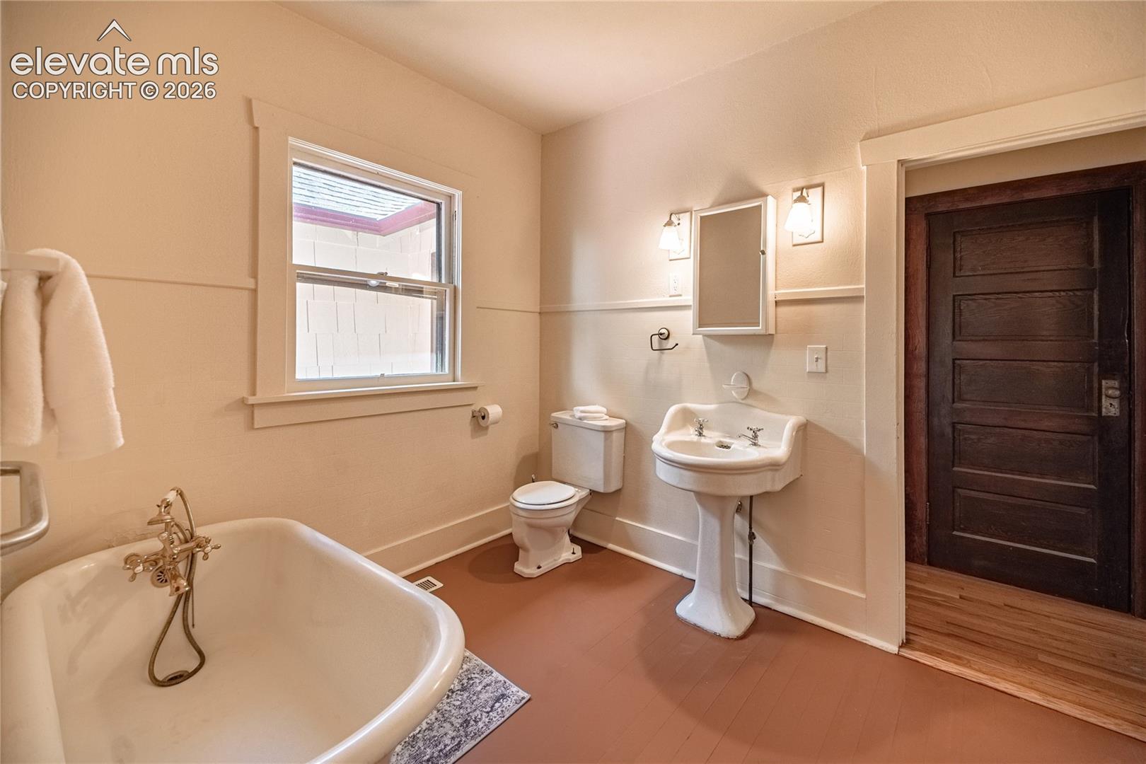Pedestal sink, clawfoot tub with brass fixtures, wood-paneled door, and a window with white trim