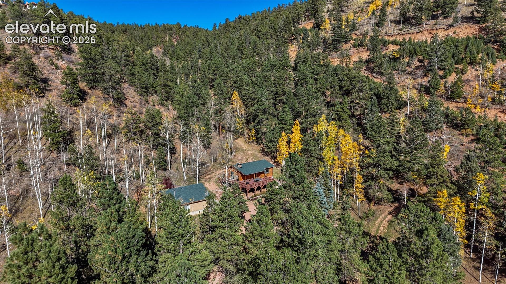 Aerial perspective of a multi-structure property featuring dark green metal roofing, elevated wooden decking, and mature coniferous and deciduous forest surroundings set against a mountainous landscape