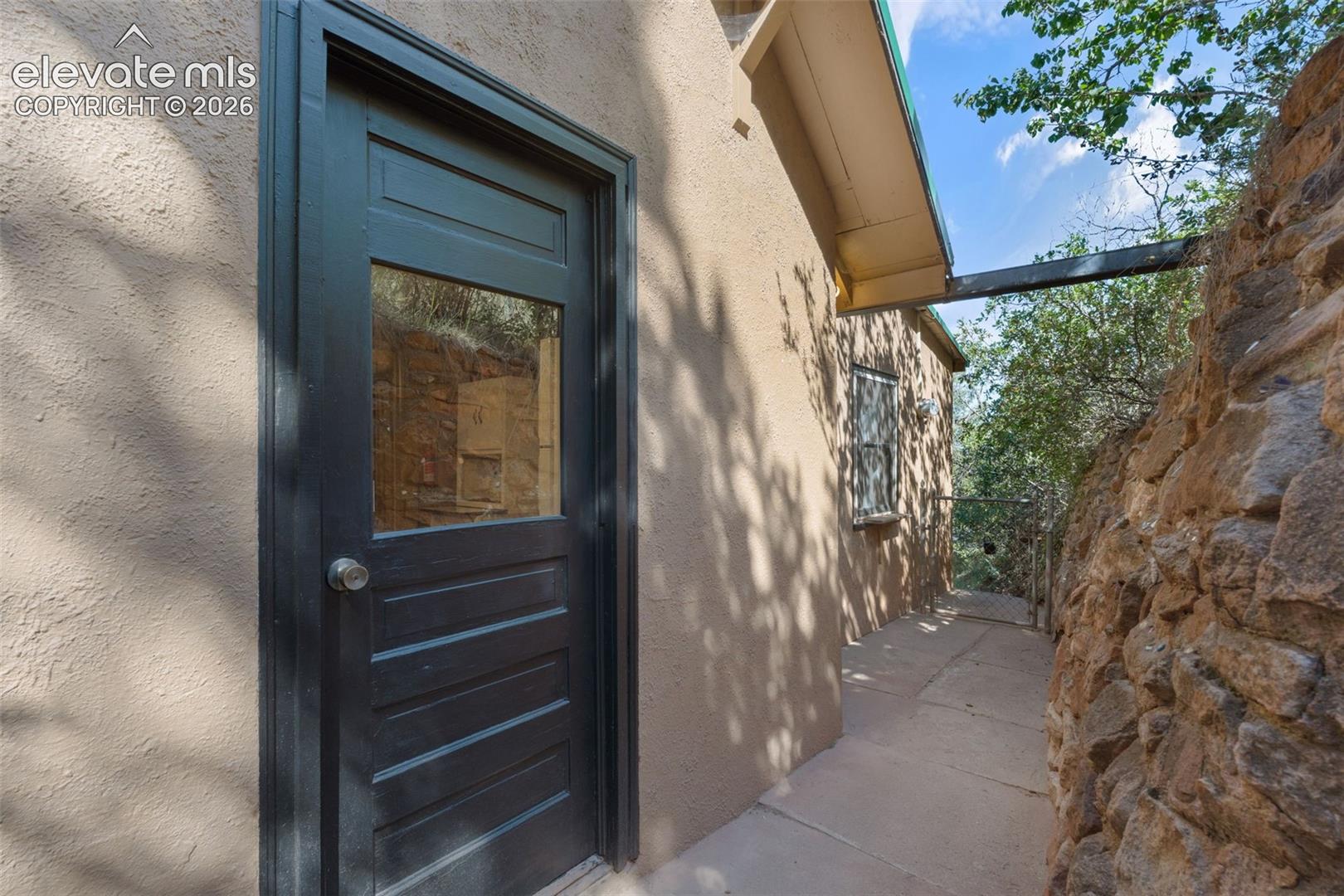 Terracotta stucco exterior featuring a dark paneled door with glass insert, a narrow concrete pathway, and a natural stone retaining wall