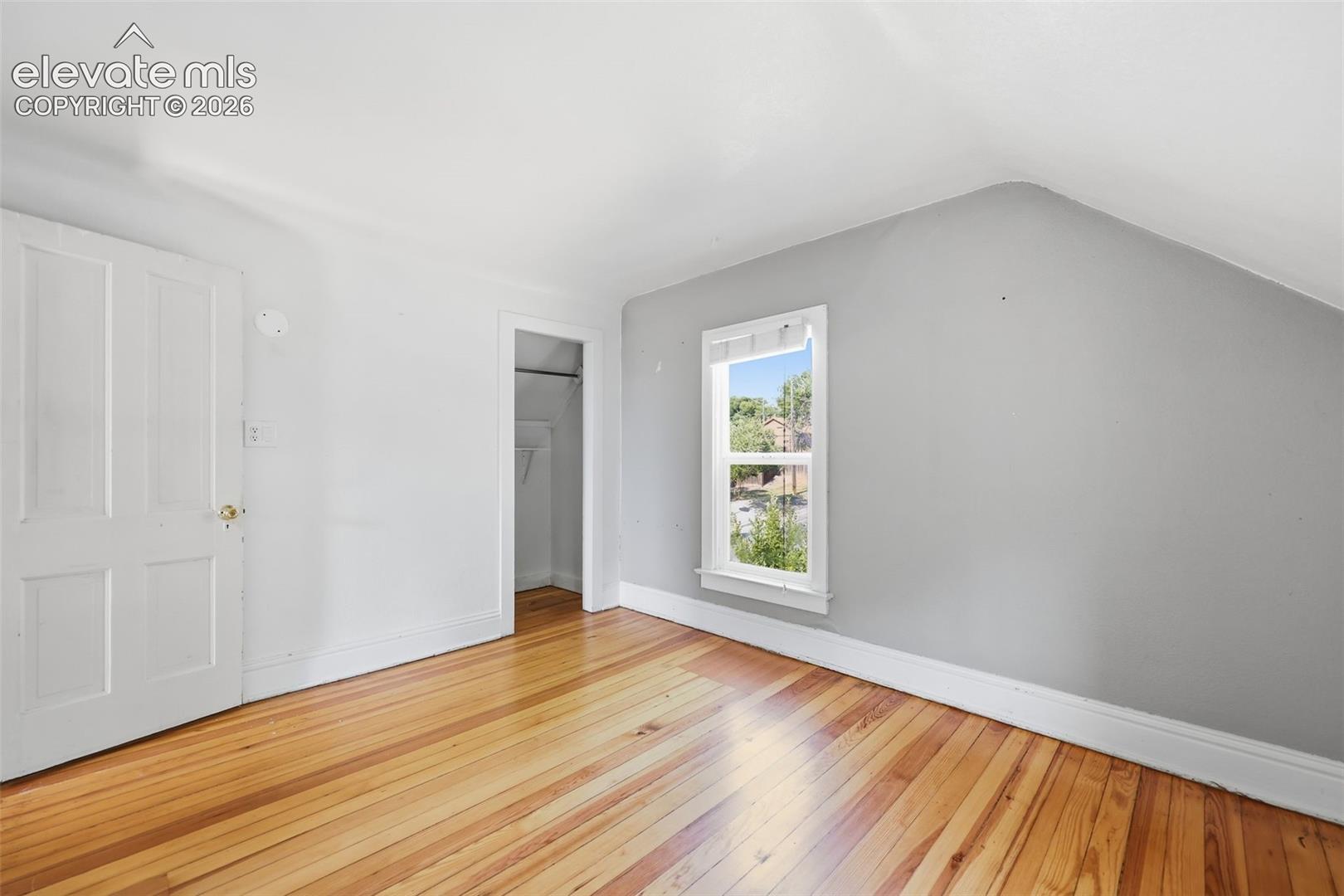 Secondary upper bedroom with pine hardwood flooring, white paneled door, sloped ceiling, and a large single window.