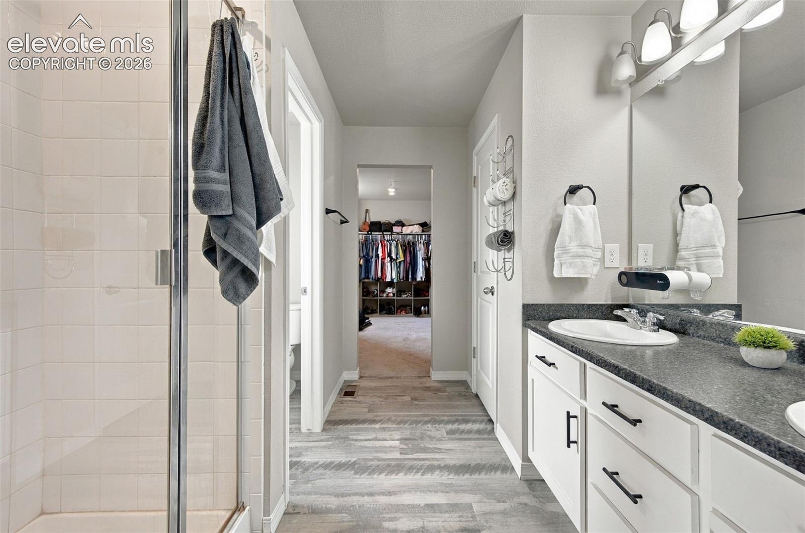 Bathroom featuring a glass-enclosed shower with white wall tile, a vanity with a dark countertop and integrated sink, white cabinetry with matte black hardware, and wood-finish flooring
