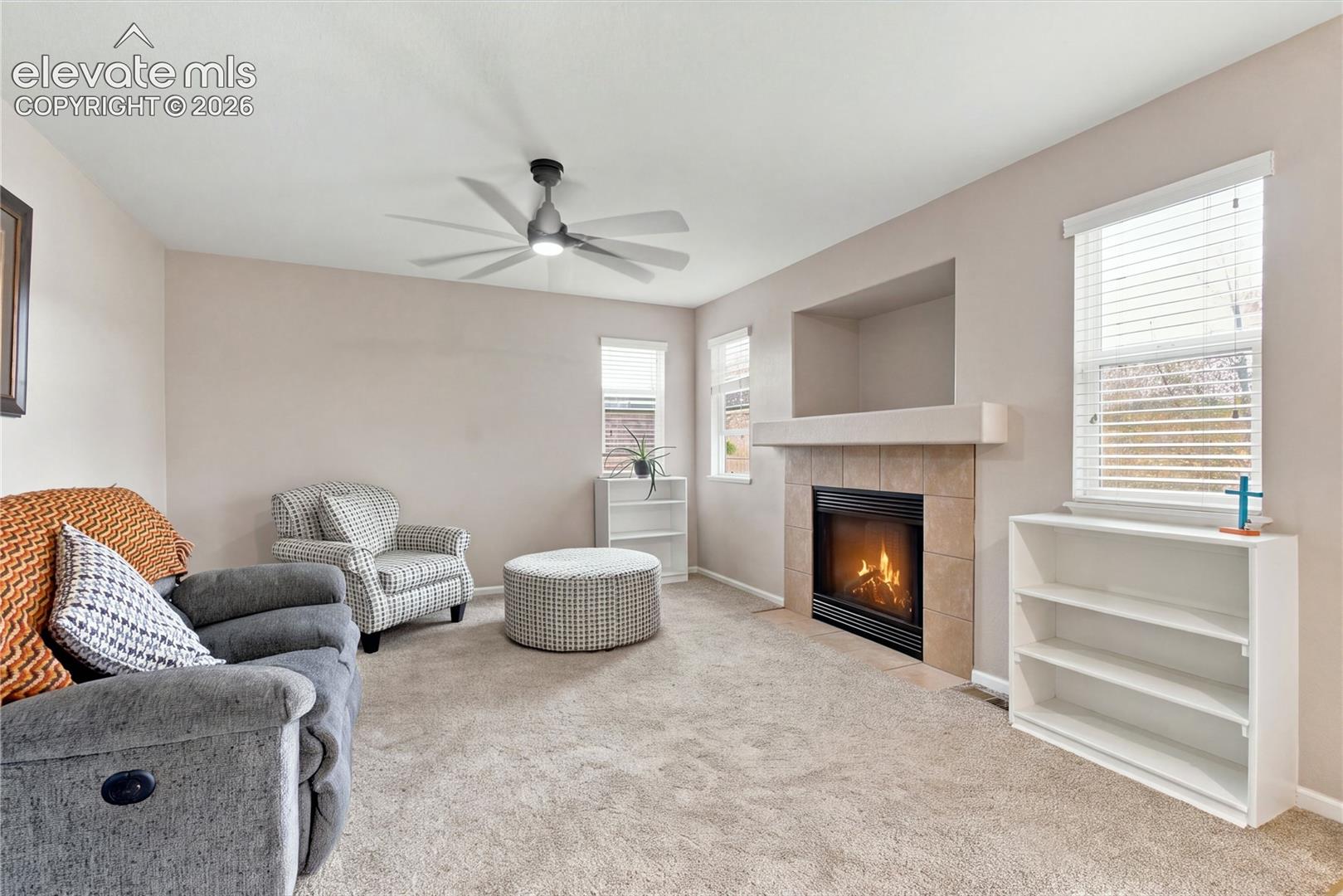Living area featuring a fireplace with a tile surround and mantel, built-in recessed wall niche, ceiling fan, and windows with horizontal blinds