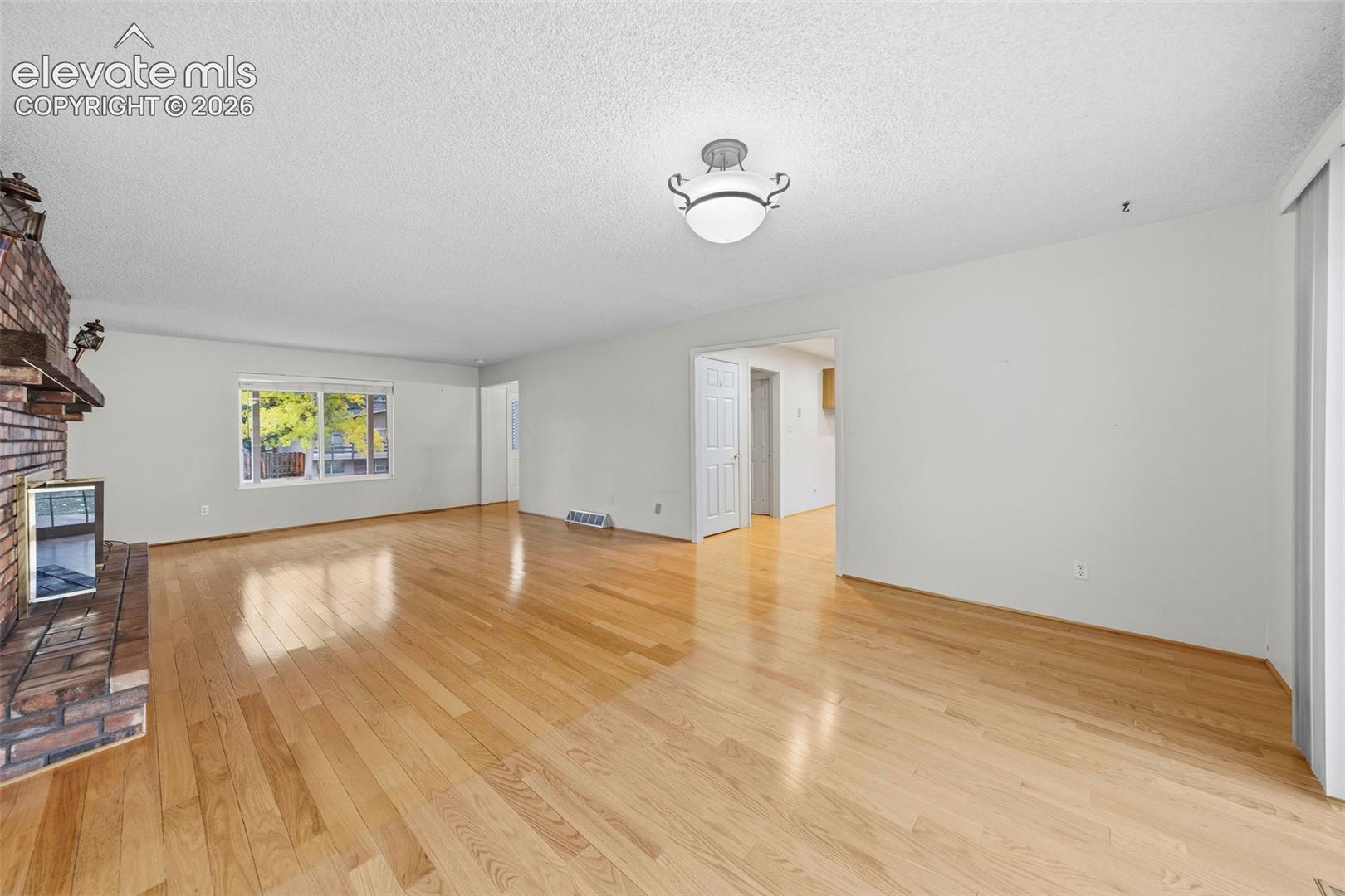Living area featuring wood-finish flooring, a floor-to-ceiling brick fireplace with a timber mantel, a recessed glass-front insert, and a flush-mount ceiling light fixture