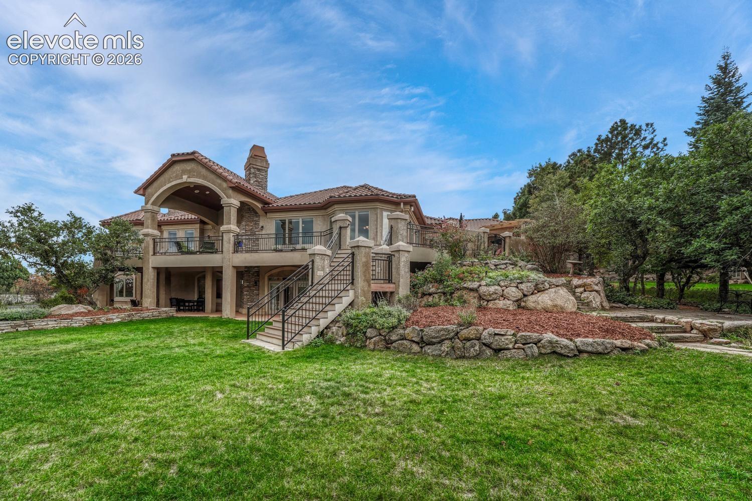 Multi-level exterior featuring stucco and stone masonry, tile roofing, an expansive elevated balcony, and tiered natural stone retaining walls