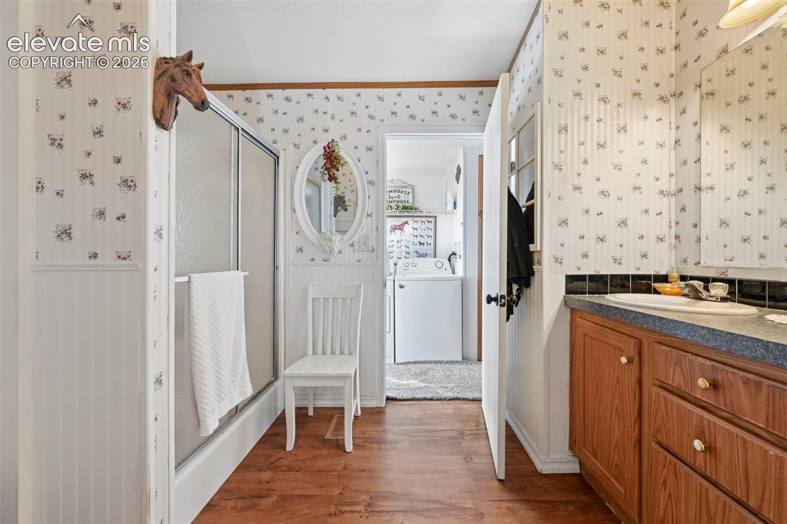 Bathroom featuring wood-finish flooring, a wood vanity with a stone-finish countertop and integrated sinks, a glass-enclosed shower, and beadboard wainscoting
