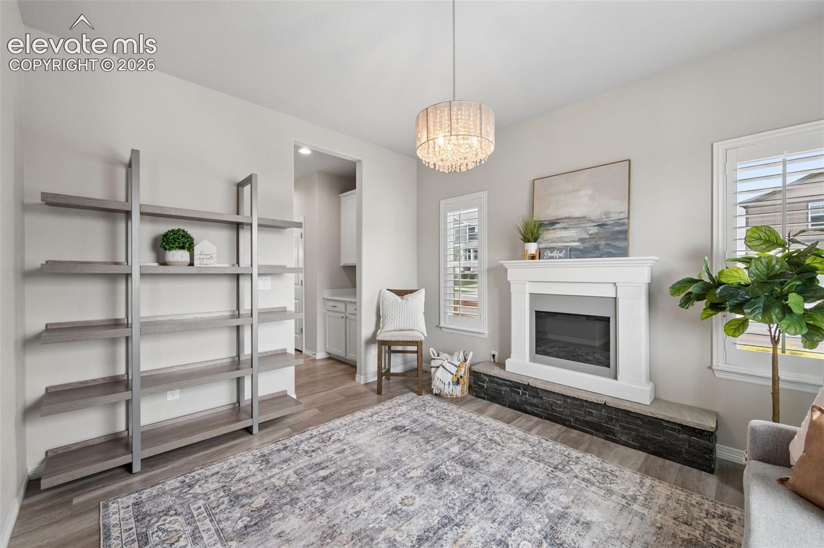 Contemporary fireplace with white mantel and dark stone hearth, wood-finish flooring, built-in shelving, chandelier, and white window shutters