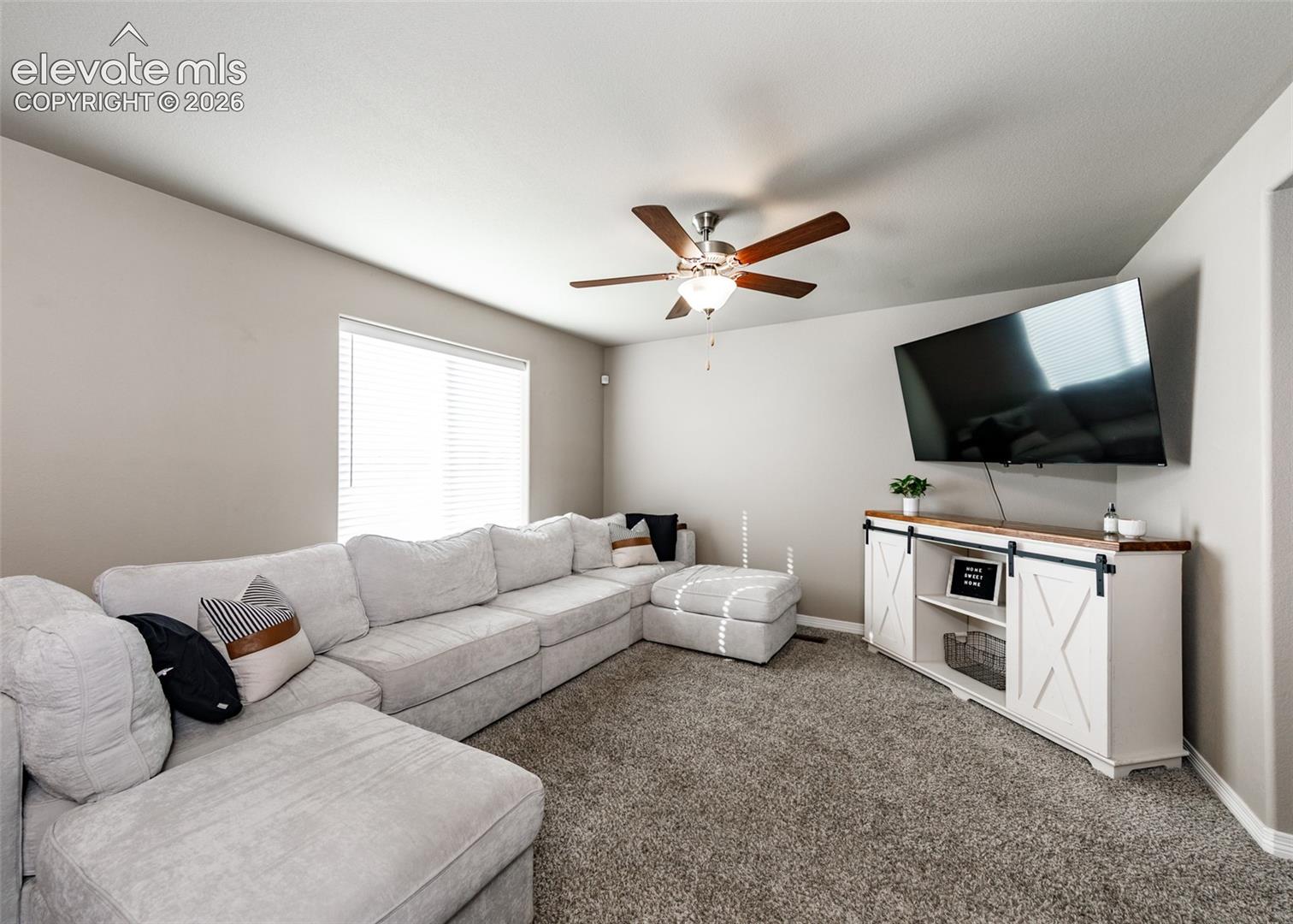 Neutral-toned living space featuring wall-to-wall carpeting, a multi-blade ceiling fan with integrated lighting, and a single-pane window with white horizontal blinds