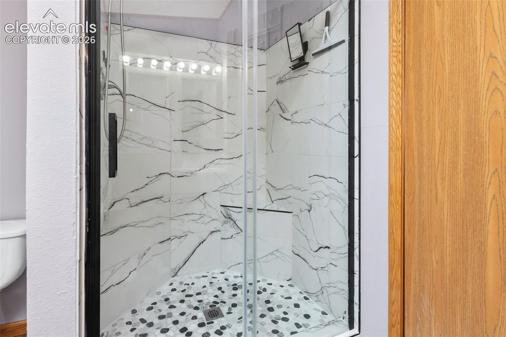 Walk-in shower featuring clear glass enclosure with dark trim, marble-patterned wall tiles, and a pebble-style floor